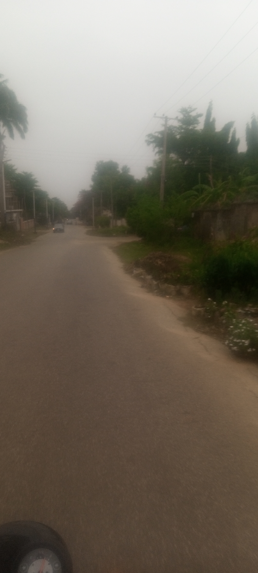 The image shows a hazy street scene in Unguwar Rimi, Nigeria. A gray asphalt road stretches into the distance, lined with green trees and vegetation on either side. Buildings are visible on the left side of the road. Utility poles with wires run along the street. A car is visible in the distance on the road. The bottom of the image shows the dashboard or instrument panel of a vehicle.