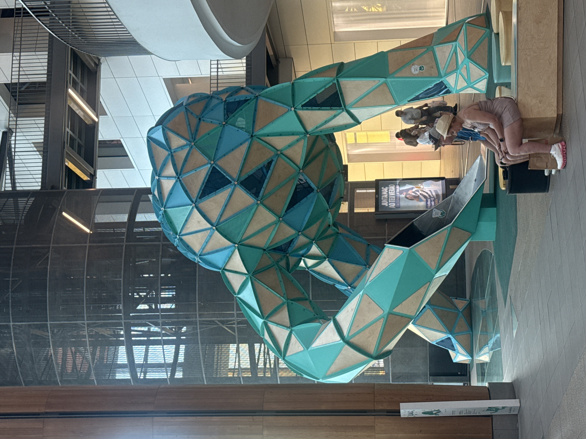 This image depicts the exterior of the Alvalade XXI shopping center, located in Porto, Portugal. The central focus is a large, abstract sculpture composed of interconnected geometric shapes in shades of teal and tan, resembling a large, segmented figure. The sculpture is positioned within an atrium or open area of the shopping center, with a multi-story structure visible in the background, featuring balconies and walkways. Natural light streams in from above, illuminating the scene.
In the foreground, two individuals are seated on a bench. The person on the right is wearing a light-colored hat and a sleeveless dress, and is looking towards the left. The person on the left, wearing a blue shirt and a dark cap, is looking straight ahead. Both are positioned in front of the sculpture. A shopping bag rests on the floor beneath the bench.
To the left of the sculpture, a tall, narrow display board with graphics and text is positioned near a thick, cylindrical column. In the middle ground, a few other people are walking or standing, some appearing to be shopping or browsing. The floor is tiled with a light-colored material in the foreground, transitioning to a patterned surface beneath and around the sculpture. Reflections are visible on the polished floor, particularly around the base of the sculpture. The overall environment suggests a public space within a modern shopping complex.