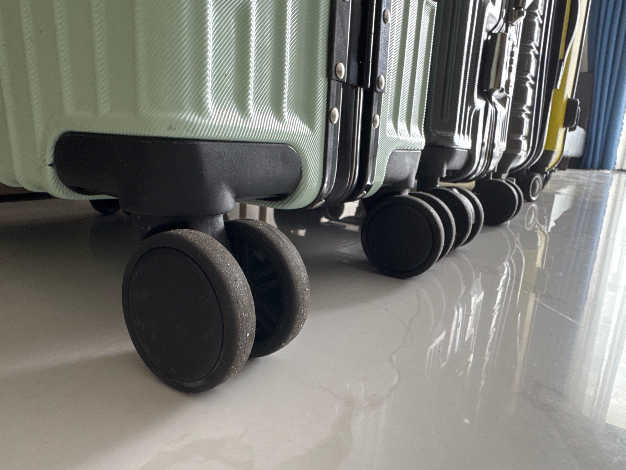 A close-up, low-angle shot captures the wheels and bottom portion of several rolling suitcases lined up. The suitcases are of various colors, including light green, dark gray, and yellow, and appear to be made of a hard shell material with ribbed detailing. They are positioned on a shiny, reflective floor, possibly tile or polished concrete. In the background, a light blue curtain is visible. The scene is indoors, with no distinct external environmental features. There are no people or animals present in the image. The overall impression is one of organized storage or display of travel luggage. The location in Nha Trang, Vietnam, is not visually discernible from the image itself.