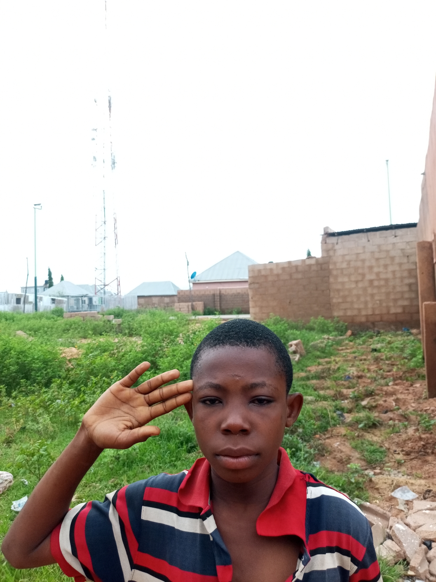 A medium shot captures a young boy in Tempure, Nigeria, saluting with his right hand raised to his forehead. He is positioned in the foreground, looking directly at the camera with a serious expression. The boy wears a striped polo shirt in red, white, and blue. Behind him, a landscape unfolds featuring overgrown green vegetation, a partially constructed brick building, and a tall telecommunications tower in the distance. The sky is overcast and bright.