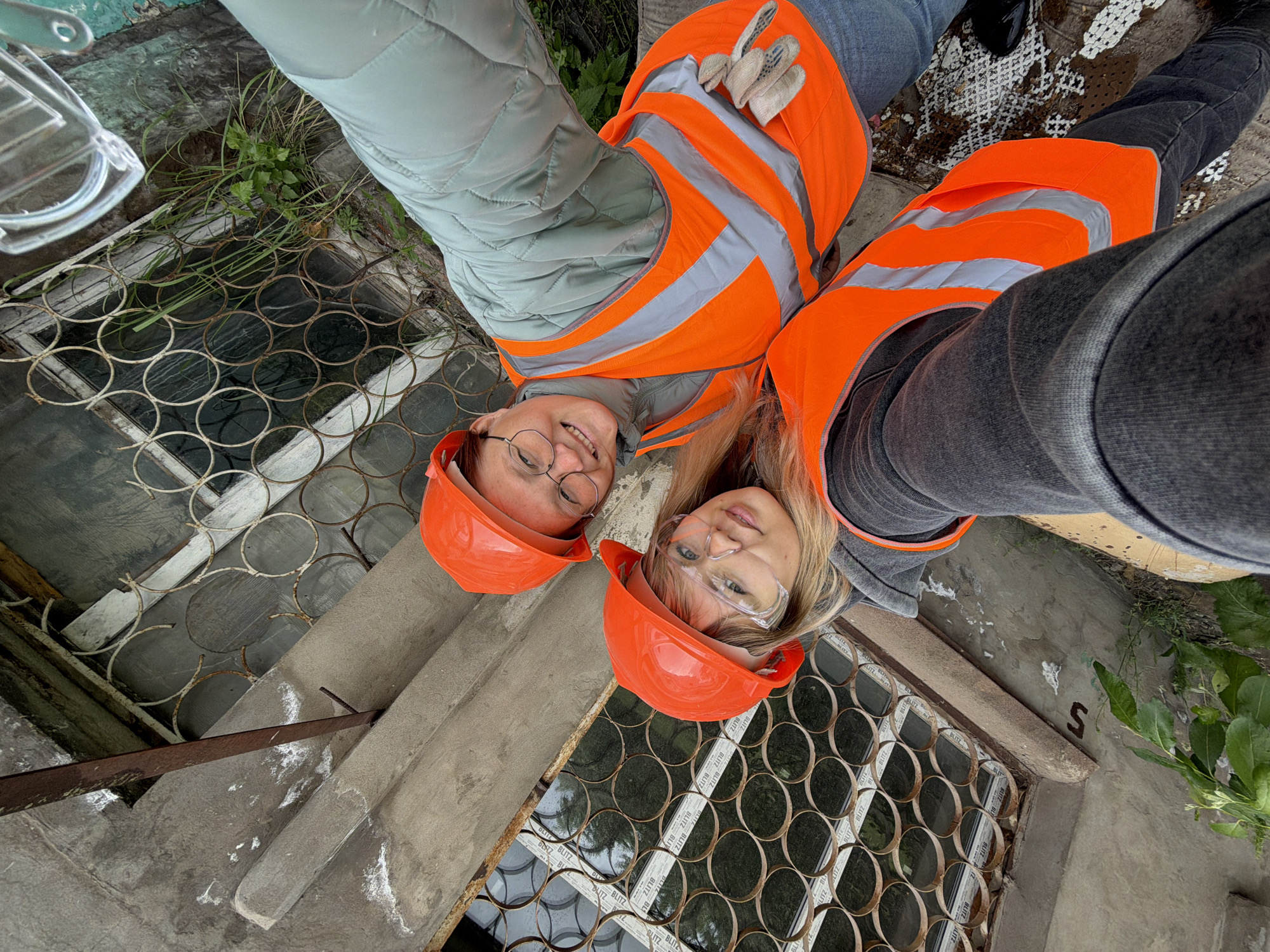 A high-angle selfie captures two smiling women dressed in safety gear at what appears to be an industrial or construction site in Chaikovsky, Russia. Both women wear bright orange safety vests with reflective stripes and matching orange hard hats. The woman on the left has light hair, round glasses, and wears a light blue quilted jacket and blue jeans. The woman on the right has blonde hair, clear safety glasses, and wears grey jeans. A pair of beige work gloves rests on the left woman's vest.
The background shows concrete structures and overgrown vegetation. Beneath the women, distinctive metal grates with a circular pattern cover what appear to be basement windows. Through these grates, white window frames are visible, bearing the repeatedly printed text "BLITZ". Other concrete surfaces show a lone letter "S" and the number "5". The scene is set during the daytime, and the women's smiles suggest a positive or friendly mood while documenting their presence at the site. A clear plastic object, possibly part of safety equipment, is visible in the top left corner.