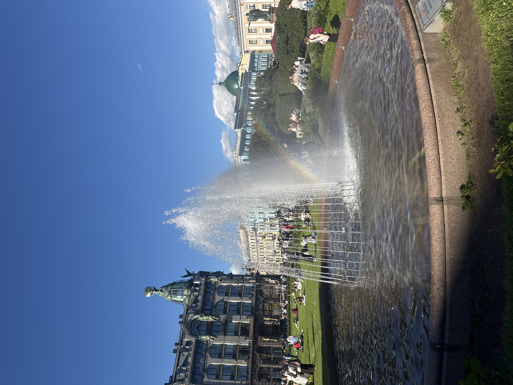 The image depicts a vibrant outdoor scene in Saint Petersburg, Russia, with a prominent fountain in the foreground spraying water upwards, creating a misty effect. A faint rainbow is visible within the spray, indicating the presence of sunlight. Several people are scattered across a grassy area in the middle ground, some sitting or lying down, while others are standing and walking. In the background, distinctive architectural structures of Saint Petersburg are visible, including an ornate building with a green dome topped by a globe and statue, and other multi-story buildings with classical facades in various colors. The sky is a clear blue with a few scattered clouds. The overall atmosphere appears to be one of a pleasant day in a public urban space.