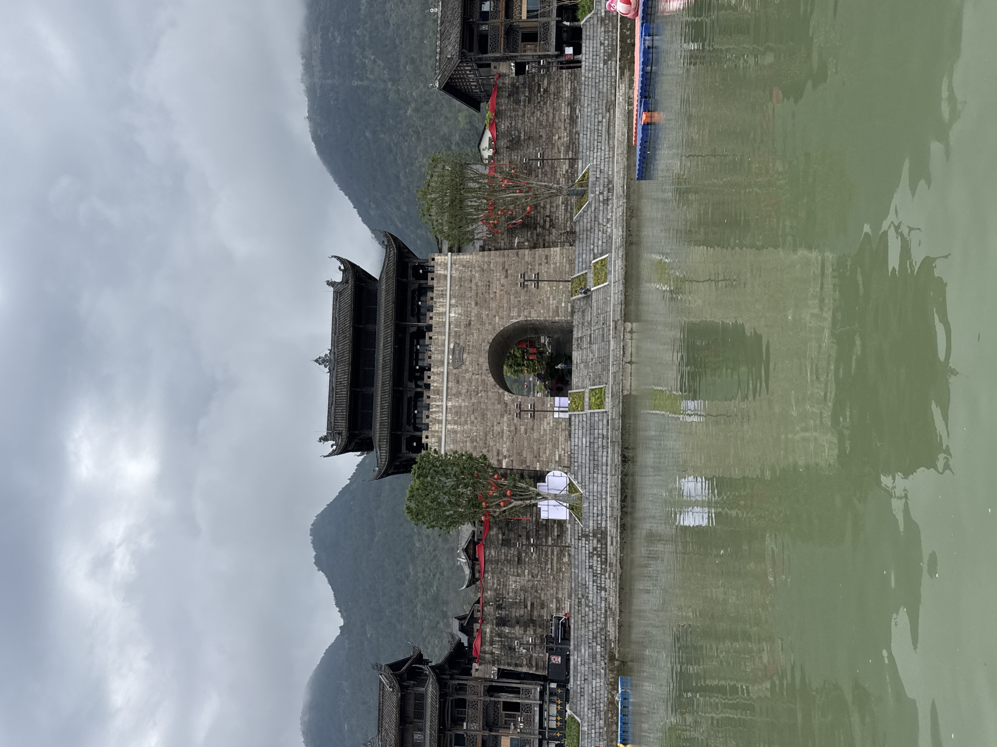The image depicts a traditional Chinese architectural scene located in Zhuoshui, China. A stone archway, possibly a city gate, dominates the center of the frame. The archway is flanked by traditional wooden buildings with ornate roofs. A mountain range rises in the background, shrouded in mist. A body of water, likely a river or lake, is in the foreground, reflecting the buildings and archway. Small trees are planted near the archway, adorned with red decorations. The sky is overcast, contributing to a subdued and atmospheric feel.