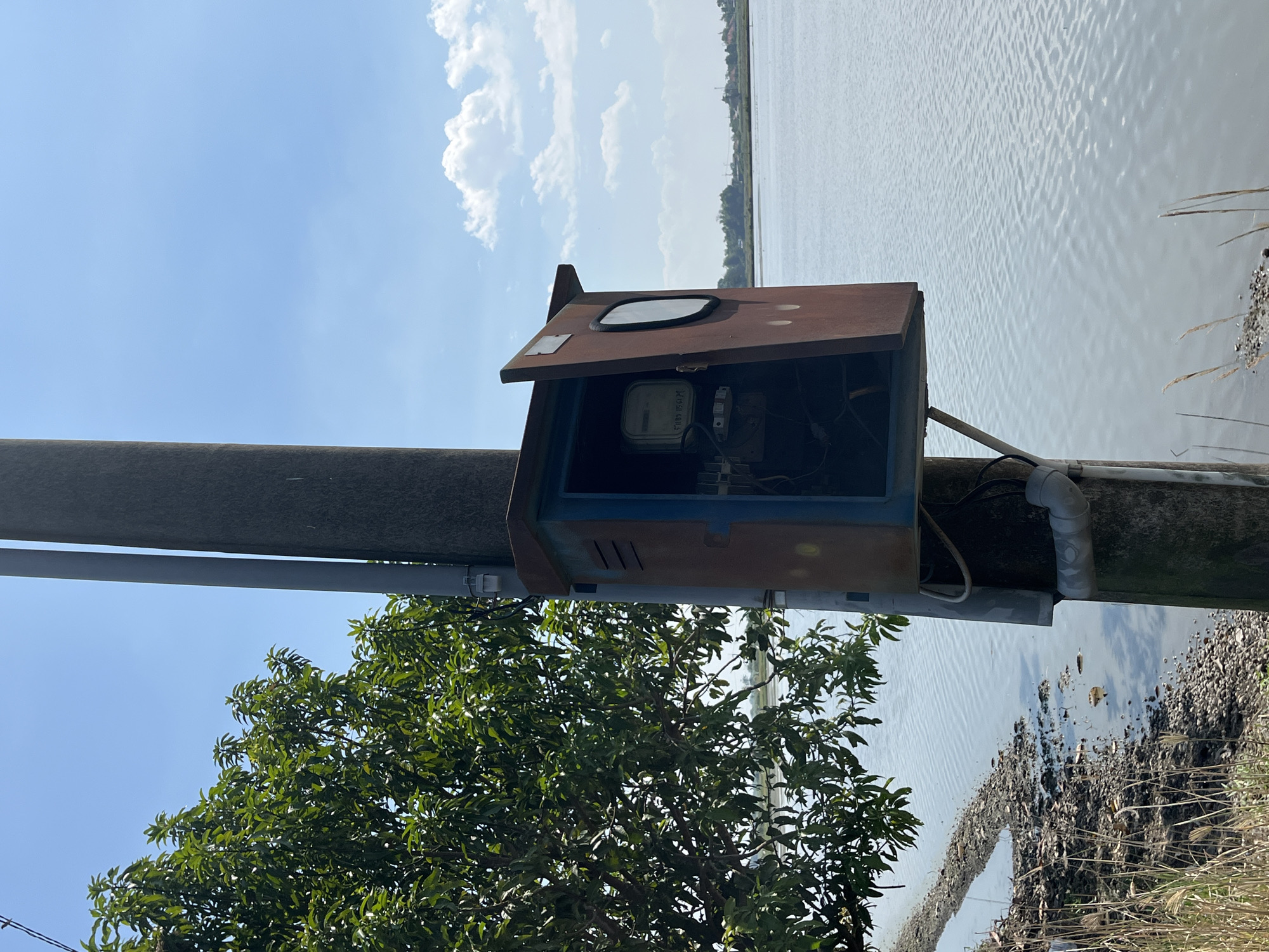 A weathered electrical junction box is mounted on a concrete pole adjacent to a body of water. The box, which appears to be made of rusted metal, has its front panel open, revealing an electrical meter inside. A large tree with green foliage is to the left of the pole, and the water extends to the horizon where a distant treeline and structures are visible under a clear blue sky with scattered white clouds. The scene is likely in or around Pesawahan, Indonesia.