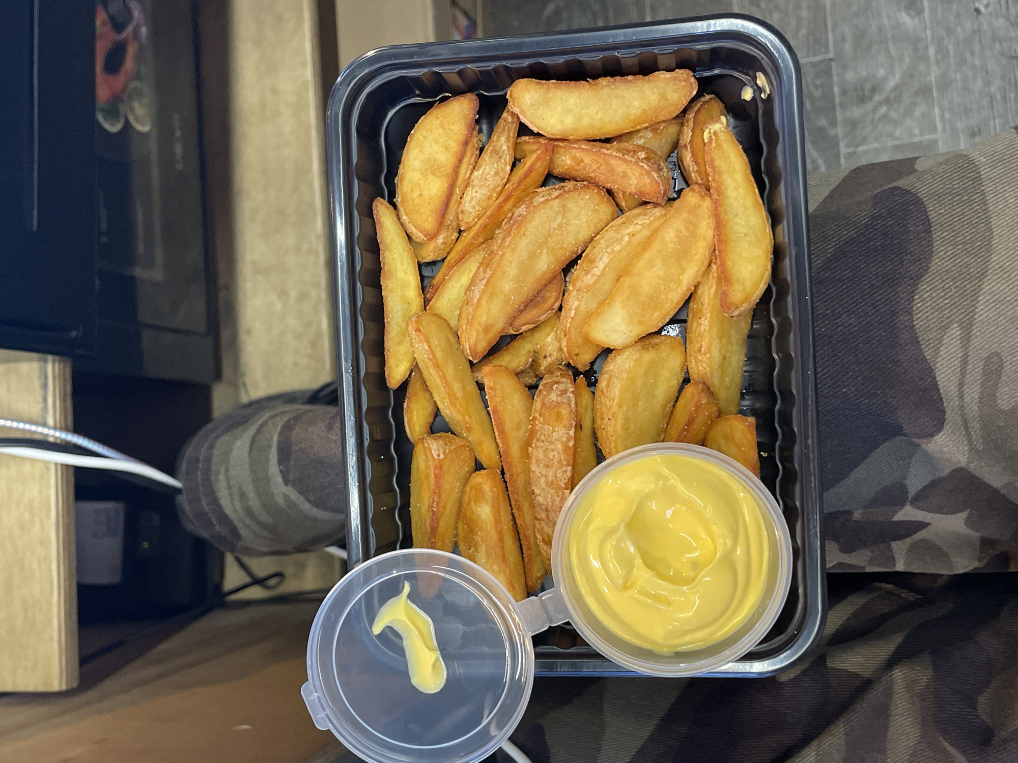 A close-up overhead shot shows a black plastic container filled with golden-brown potato wedges, some with a light dusting of seasoning. To the left, a clear plastic dipping container is open, with a small dollop of yellow sauce on the lid and a generous amount of sauce within the container. The food is placed on a surface with camouflage-patterned fabric underneath. In the background, out of focus, a wooden surface and electronic equipment are visible. The context of Shiryaevo, Russia, is not evident in the image.
