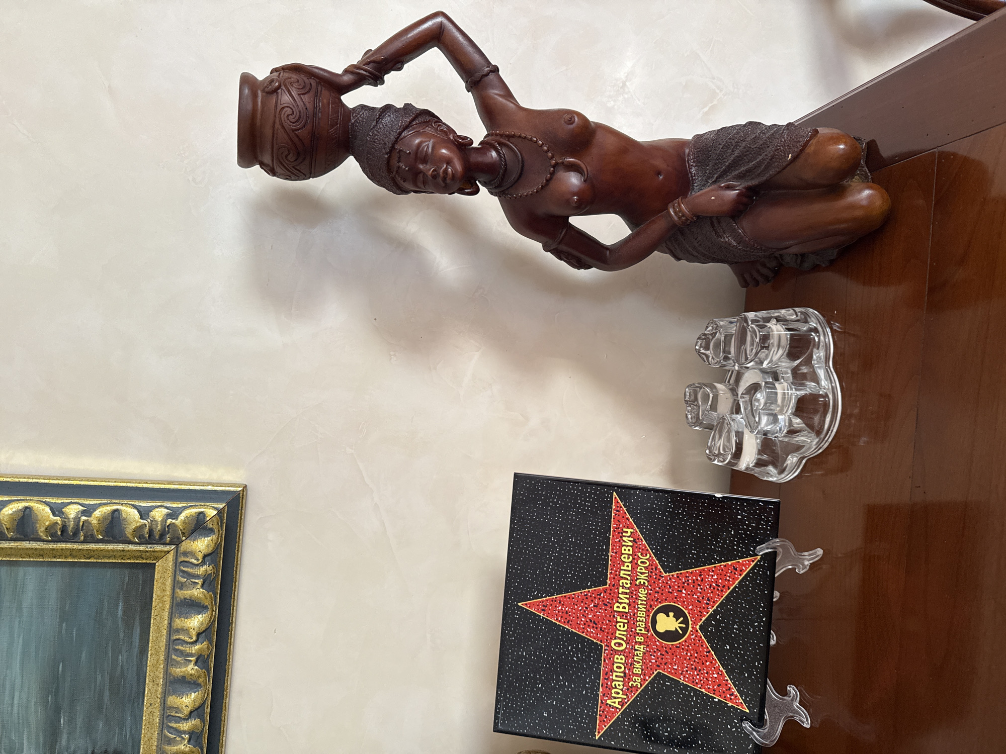 In the city of Yukki, Russia, on a polished wooden surface, a dark book with a red star design is displayed on a clear acrylic stand. The book features text in yellow and white, with the prominent text reading "Арапов Олег Витальевич" and "За вклад в развитие ЭКРОС", along with a circular emblem containing a film camera. To the right of the book, a wooden figurine of a kneeling woman is positioned. The figurine depicts a woman with her head tilted back, a pot balanced on her head, and wearing traditional jewelry and a wrap around her waist. The background is a textured, light-colored wall. In front of the figurine, a clear glass candle holder with three cups is visible. The scene appears to be an indoor setting, possibly a home or an office, with the lighting casting shadows on the wall.