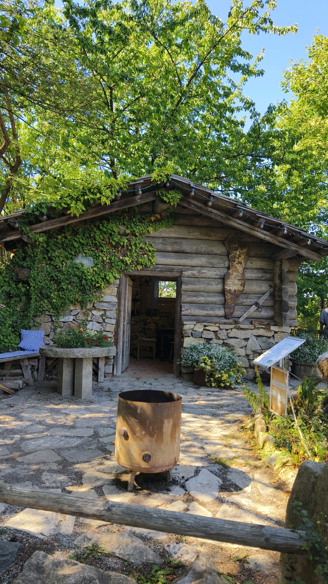 The image depicts the exterior of a rustic log cabin and stone structure, likely part of an outdoor exhibit or historical site, situated in Langenlois, Austria. The cabin, constructed from horizontal logs, has a stone-foundation and is partially covered by dense green ivy. A wooden roof with decorative lights overhangs the entrance, which is open, revealing a dimly lit interior with visible furniture and a window. In front of the cabin, a stone table with a checkered cushion on a wooden bench is visible. A large, rusty cylindrical object on legs, resembling an old furnace or incinerator, stands in the foreground on a stone-paved courtyard. The area is surrounded by mature trees with lush green foliage, casting dappled sunlight on the ground. Other elements include potted plants, a sign, and rough-hewn wooden fencing. No people are clearly visible in the image.