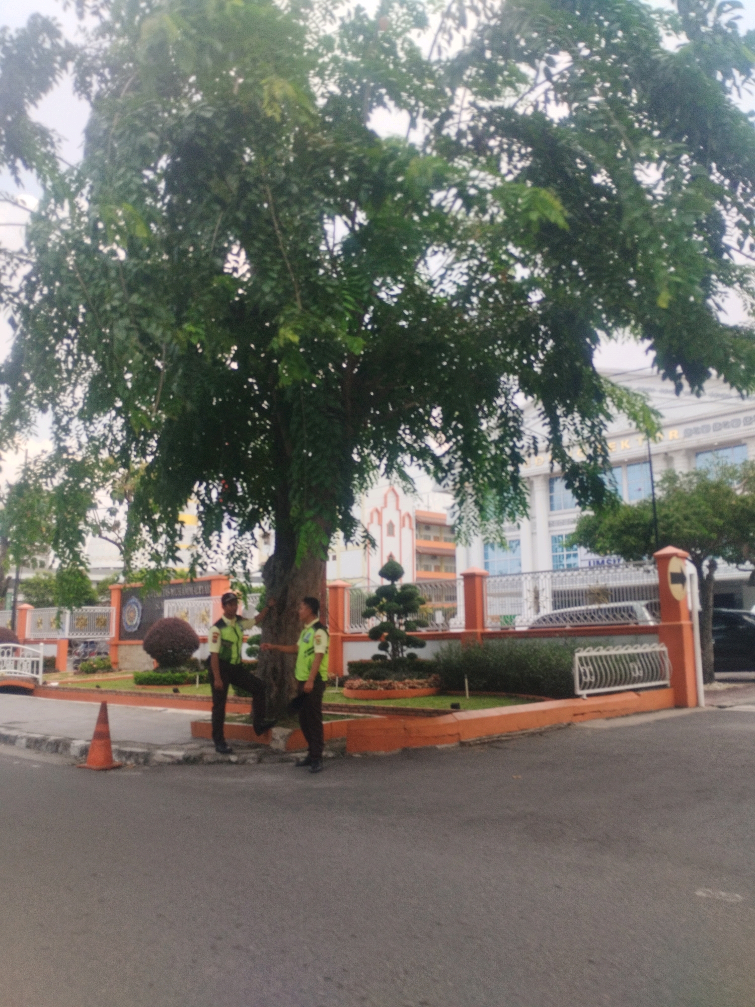 Two security guards in bright green vests stand by a large tree in front of a building. The building appears to be a white, multi-story structure with several windows and architectural details. In the foreground, there is a paved road and a concrete curb. A traffic cone is positioned on the curb. The area also features an orange-colored wall and a white metal fence, with some greenery and a topiary plant. The sky is overcast. The image was captured in the City of Medan, Indonesia.