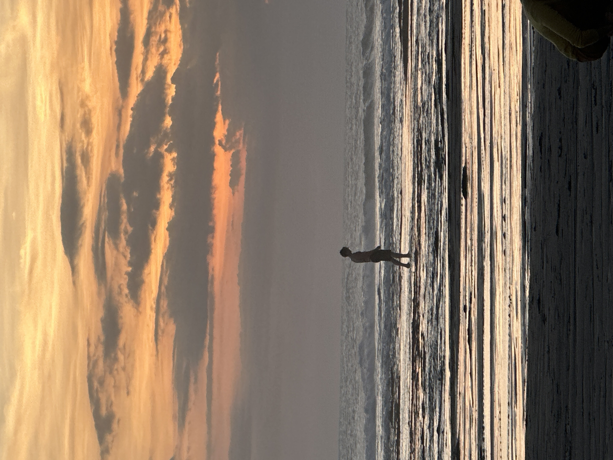 A slightly overexposed landscape photograph depicts a beach scene. The image is divided into three horizontal sections. The left third of the image shows an orange and yellow sky with layers of striated clouds. The middle third of the image shows the grey tones of the ocean meeting the sky, with waves visible near the horizon. A single figure is silhouetted in the ocean. The right third of the image appears to be a darkened section of sand or other land with a ribbed or striated pattern, with a hint of a green or yellow object in the top right corner. The location is in the city of Mengwi, Indonesia.