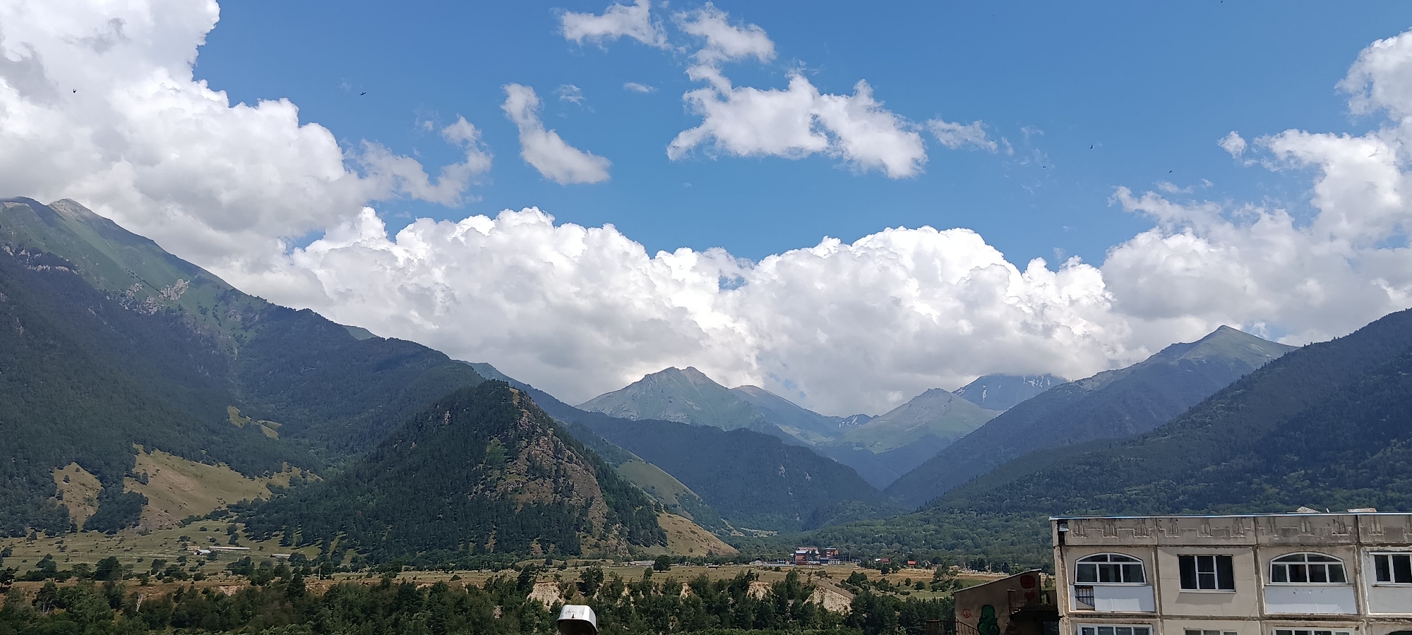 The image captures a panoramic view of a mountainous landscape under a partly cloudy blue sky. The foreground features a multi-story building with a light-colored facade and numerous windows. Beyond the building, the terrain slopes upward, covered in lush green forests that transition into the rugged slopes of a mountain range. The mountains are layered, with the closest ones densely forested and the distant peaks showing bare rock and patches of snow. The sky is dotted with white, puffy clouds, and two small dark objects, possibly birds or aircraft, are visible in the upper sky. The scene appears to be a scenic overlook in the city of Teberda, Russia, showcasing the natural beauty of the region.