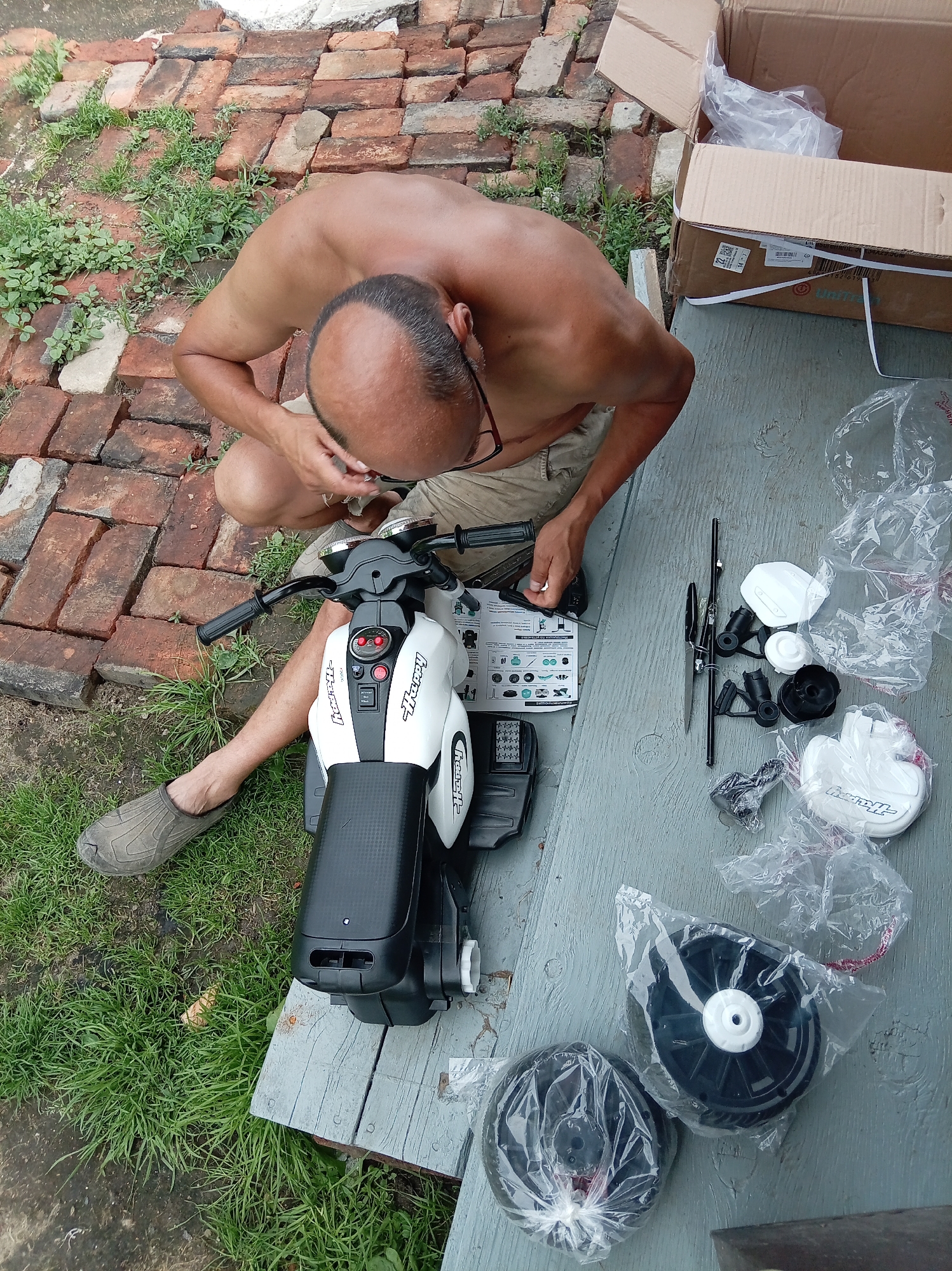 The image shows a shirtless, balding man with glasses assembling a white and black toy motorcycle outdoors. He is kneeling on grass next to a pile of red bricks and a wooden surface. Scattered around him are various parts of the toy motorcycle, including wheels, plastic casings, and instruction manuals, some still in plastic packaging. A large cardboard box, likely containing the toy, is visible in the background. The scene appears to be a residential backyard or outdoor workspace. The specific location in Artyom, Russia, cannot be determined from the image alone.