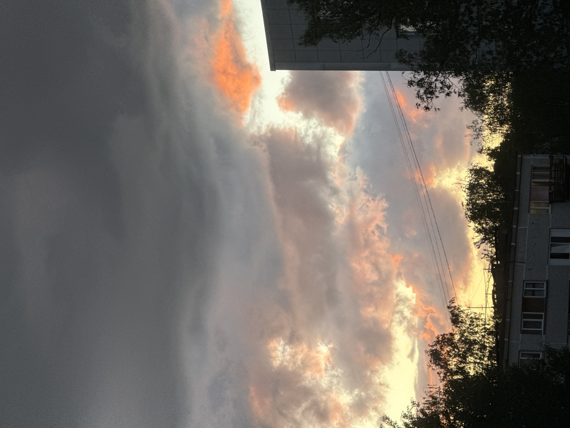 The image depicts a cloudy sky in what appears to be an urban environment. Orange hues are visible in the clouds, suggesting a sunset or sunrise. A building is partially visible at the top and on the side of the frame, indicating the picture was taken from a street or residential area. Some trees are also visible in the foreground. It is not possible to confirm the exact location in Riga, Latvia from this image alone. No people, subjects, objects, or actions are visible beyond the buildings, trees, and sky.
