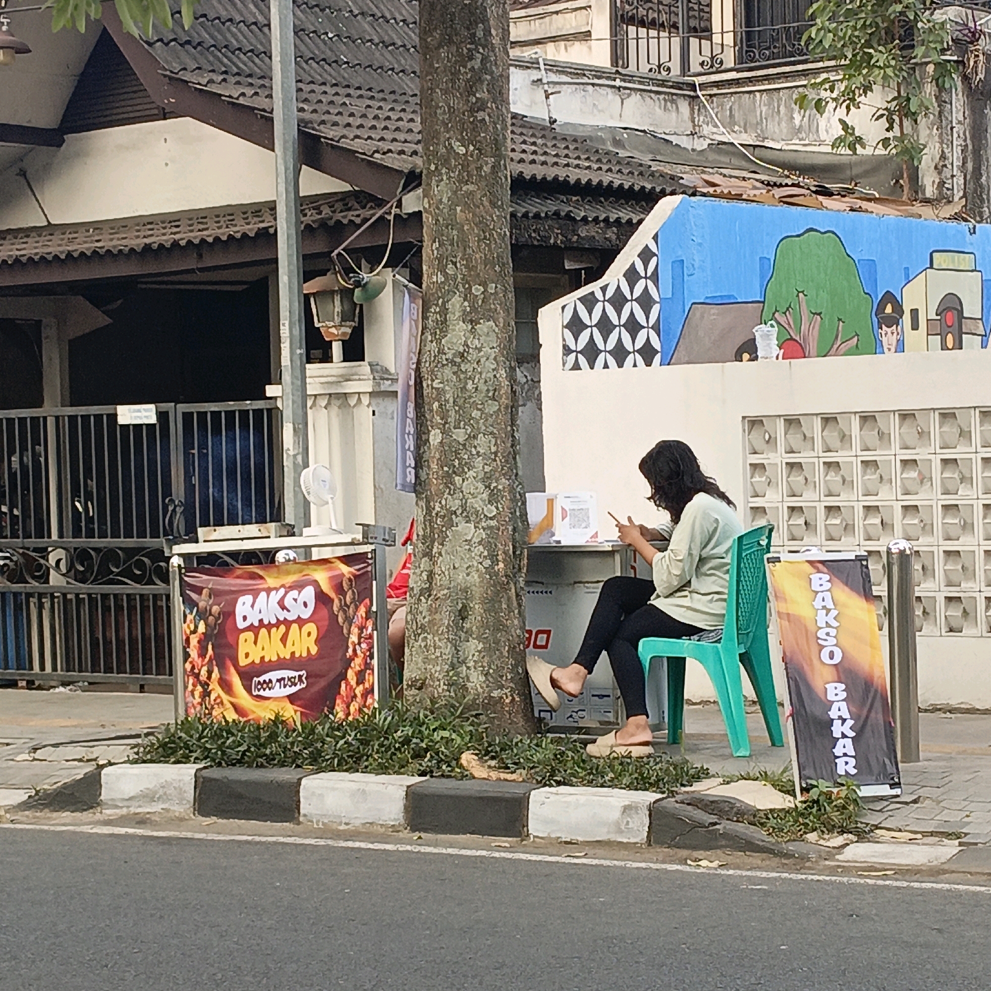 The image depicts a street scene in Salatiga, Indonesia, where a woman is sitting on a bright green plastic chair next to a tree. She is wearing a light-colored long-sleeved shirt and black leggings, with beige slip-on shoes. The woman is looking down at her smartphone, which she is holding in her hands.
In front of her, on the sidewalk, there is a small food stand or cart. Two banners are displayed at the stand. One banner, on the left, is dark red with text that reads "BAKSO BAKAR" in large yellow letters, accompanied by images of grilled meat skewers and flames. The other banner, on the right, is also yellow and orange and appears to have vertical text that also says "BAKSO BAKAR".
Behind the woman and the stand is a white wall with a colorful mural. The mural features a blue sky, a large green tree, a police booth with a traffic light, and a drawing of a person in a police uniform. The wall has decorative white patterns on the left side. A grey metal fence with decorative scrollwork is visible behind the stand and to the left. A large tree trunk stands prominently in the foreground, partially obscuring the view of the food stand and the woman.
The scene is on a paved sidewalk next to a road. The ground is covered with concrete and there is a section of grass and small plants between the sidewalk and the curb. The overall impression is of a casual outdoor setting, possibly a street food vendor operating in a residential or commercial area.