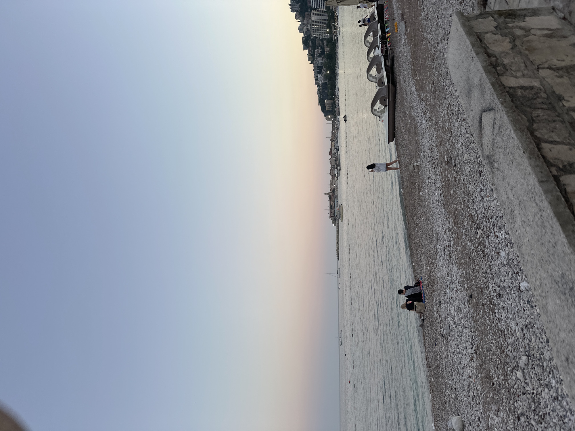 The image depicts a serene dusk scene at a pebble beach in Budva, Montenegro. In the foreground, a stone wall is visible, with a section of the pebble beach extending towards the sea. Several paddle boats are lined up on the beach, some with people interacting around them. Further down the beach, a couple is seated on the pebbles, facing the water, with a bag placed beside them.
The sea stretches out towards the horizon, with a few boats visible on the water, including sailboats and a jet ski. In the distance, the Budva cityscape is illuminated, with buildings and structures climbing the hills overlooking the coast. The sky exhibits a gentle gradient of colors, transitioning from a pale blue at the top to soft hues of pink and orange near the horizon, indicating the time of sunset. The overall atmosphere is calm and tranquil.