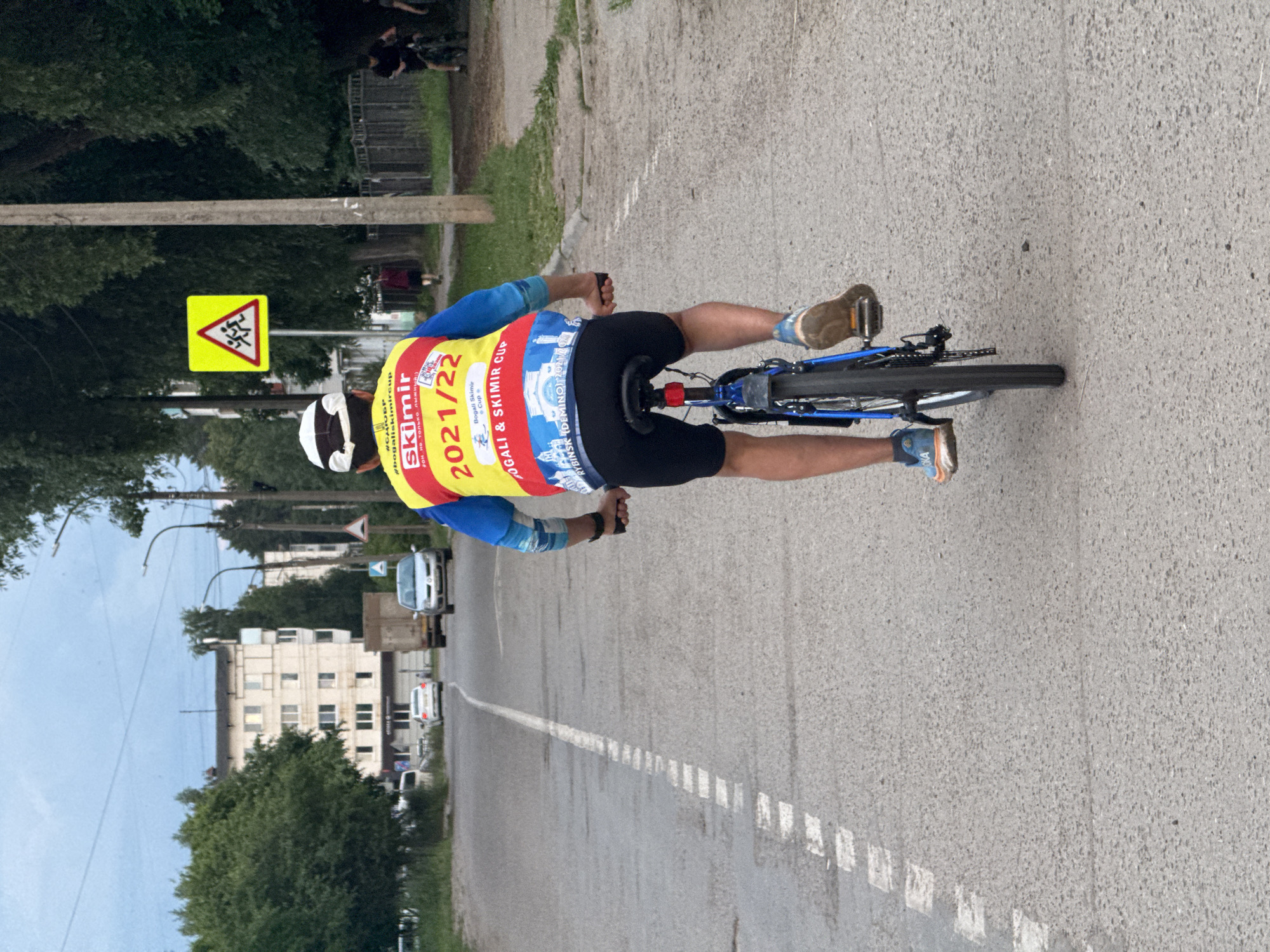 The image shows a person riding a bicycle on a paved road in Chaikovsky, Russia. The cyclist is wearing a blue and yellow long-sleeved jersey with "SKIMIR 2021/22" and other text and logos. They are also wearing black shorts, a white cap, and blue and orange athletic shoes. The bicycle is blue and has a red taillight. In the background, there are trees, buildings, streetlights, a yellow triangular road sign with figures of children, and several vehicles including cars and a truck. The road has a white dashed line marking the lane. The sky is overcast.