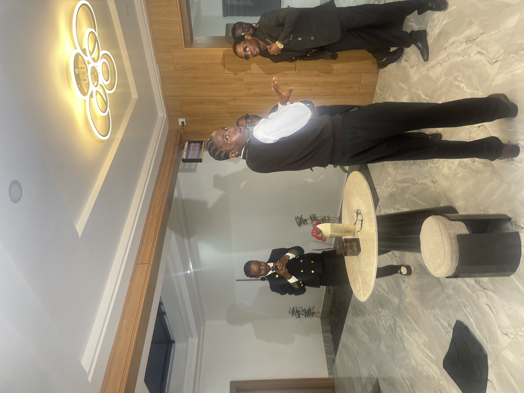 This image captures a social gathering in what appears to be a modern indoor space, potentially a hotel lobby or event venue, in Gura Top, Nigeria. The lighting suggests it is evening or a dimly lit interior. The room features a polished marble floor and a wooden-paneled wall. A circular table with a light-colored top sits in the foreground, adorned with a vase of red flowers, a decorative box, and a pair of glasses.
Several individuals are present, all appearing to be dressed in formal or semi-formal attire. One person in the foreground, with braided hair, is facing the camera and gesturing with their hands while wearing a dark vest over a white shirt. To their left, another individual in a dark suit with gold embellishments on the lapels is looking towards the foreground person. Further in the background, more individuals in suits are visible, some partially obscured. One person in the middle ground is standing near the table, looking down at their phone while wearing a dark suit jacket with gold buttons. The ceiling has a distinctive modern light fixture consisting of interlocking circular elements. The overall impression is that of a gathering or event where individuals are engaged in conversation or interaction.