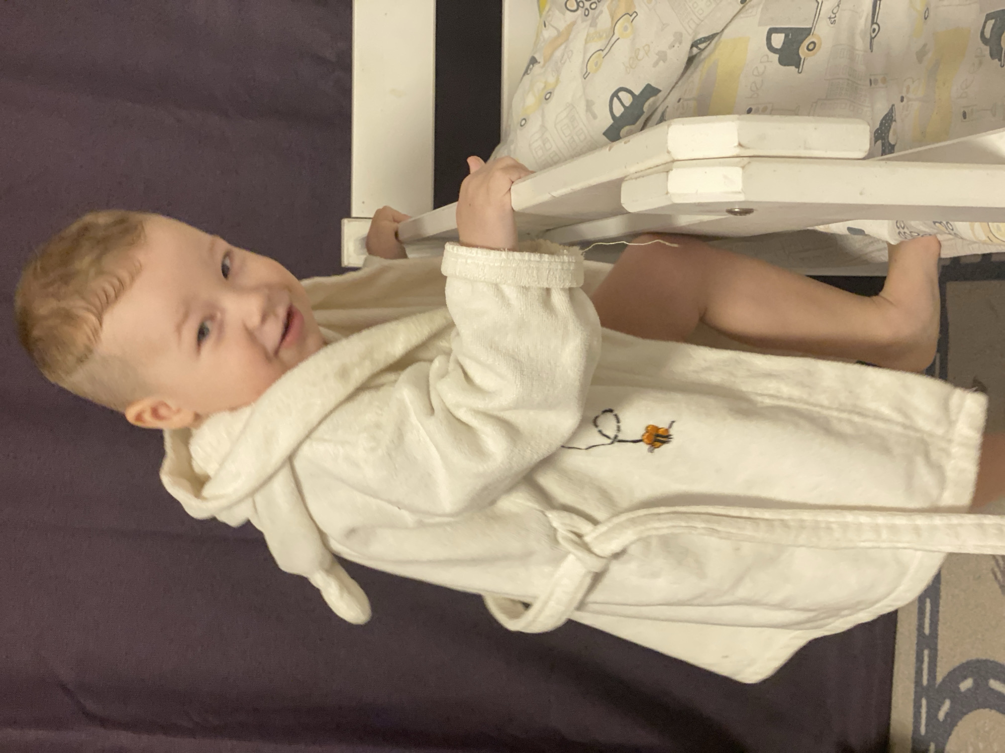 A fair-skinned baby with light hair and chubby cheeks is lying down, looking towards the viewer with a joyful smile. The baby is wearing a white, hooded bathrobe, tied at the waist, which features a small embroidered design of a bee or insect on its left chest. Both of the baby's hands are gripping the white wooden bars of a crib or playpen.
Inside the crib, patterned bedding is visible, adorned with illustrations of cartoon cars, buildings, and the word "beep". To the left of the baby, a dark purple surface, possibly a blanket or bedding, takes up a significant portion of the background. A faint patterned surface resembling a road or track can be seen beneath the baby's leg on the right side of the frame. The scene takes place indoors, likely in a nursery or bedroom in Sochi, Russia.