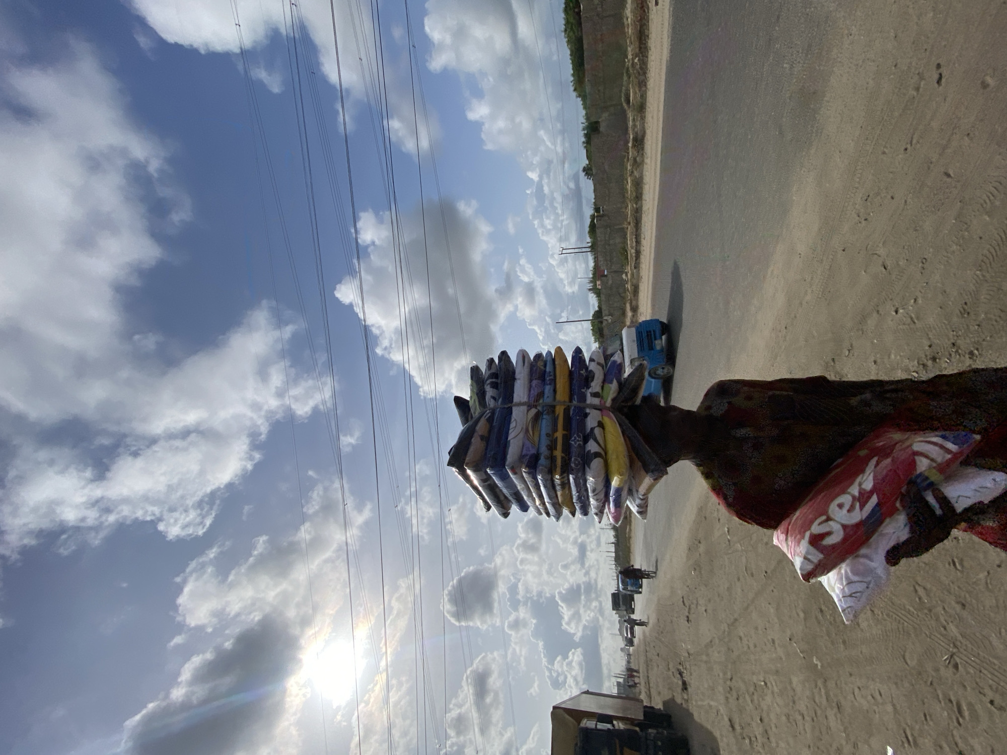 A vendor carries a stack of goods on their head along a road in Lekki, Nigeria. The goods appear to be wrapped clothing or textiles, secured with bands. The vendor is carrying an additional bag on their arm. The road is paved, with a sandy shoulder. A vehicle is parked near the shoulder and other vehicles are visible further down the road. The sky is partly cloudy, with power lines running overhead. The sun is shining brightly.