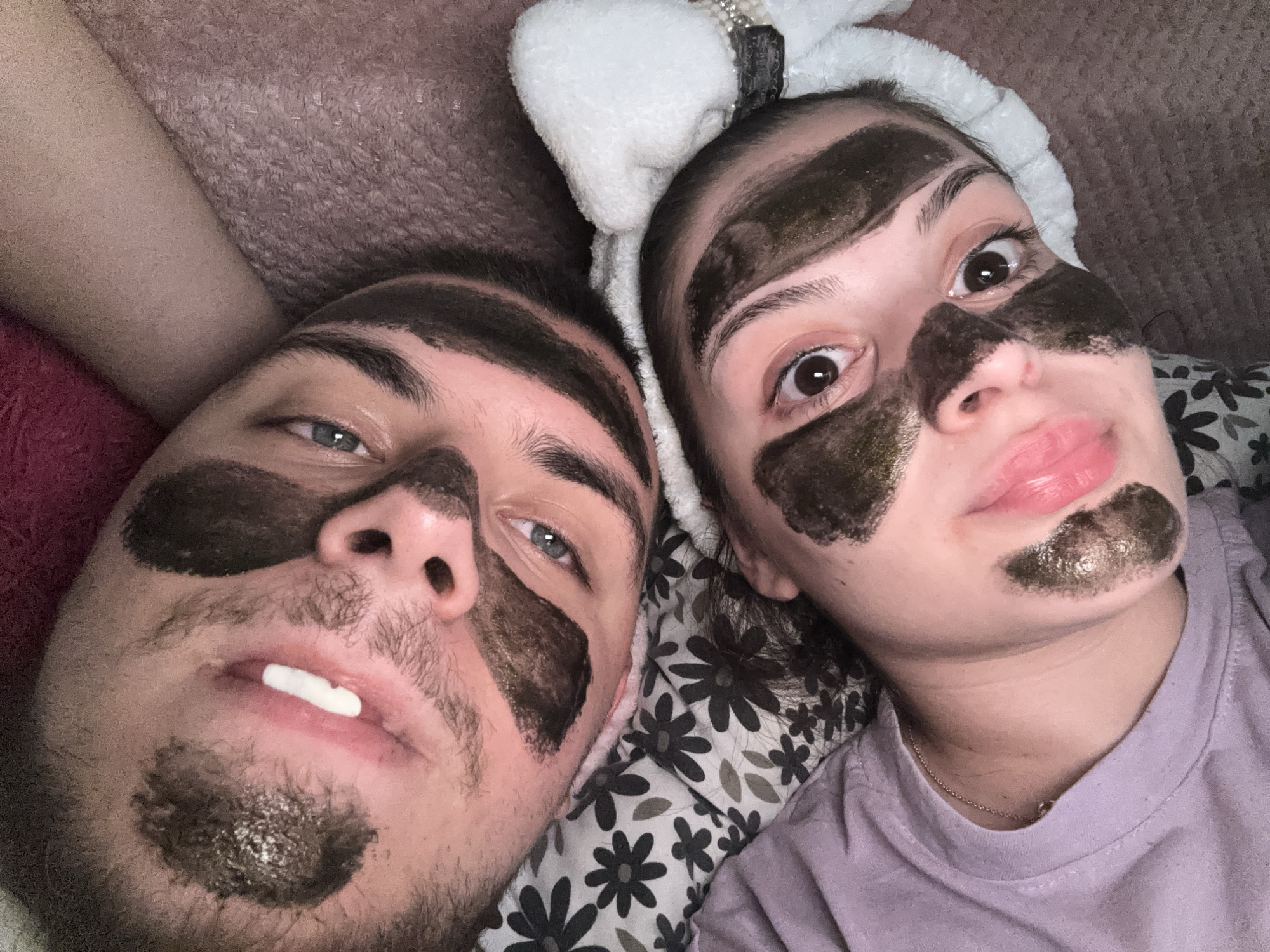 An indoor, close-up image, purportedly taken in Burgas, Bulgaria, depicts two individuals, one male and one female, lying side-by-side. Both individuals have dark, possibly charcoal, face masks applied in irregular patches across their foreheads, under their eyes, on their cheeks, and chins.
The individual positioned on the right is a female with wide-open, dark brown eyes directly engaging the camera. Her lips are a glossy pink and slightly parted in a pout. She wears a white, fluffy headband, possibly adorned with pearls, pulling her hair back from her face. She is dressed in a light purple crew-neck t-shirt and a thin gold chain necklace. Her head rests on a pillow with a black and white floral pattern.
The individual on the left is a male with light blue-green eyes, also looking directly at the camera. He exhibits stubble or short facial hair on his chin and upper lip. A small, elongated white object is visible between his slightly parted lips. He is resting on a dark red or pinkish textured blanket or sheet. Part of his arm, appearing beige, is visible to the upper left. The scene suggests a relaxed, shared activity, likely within a bedroom setting, under soft, ambient lighting.