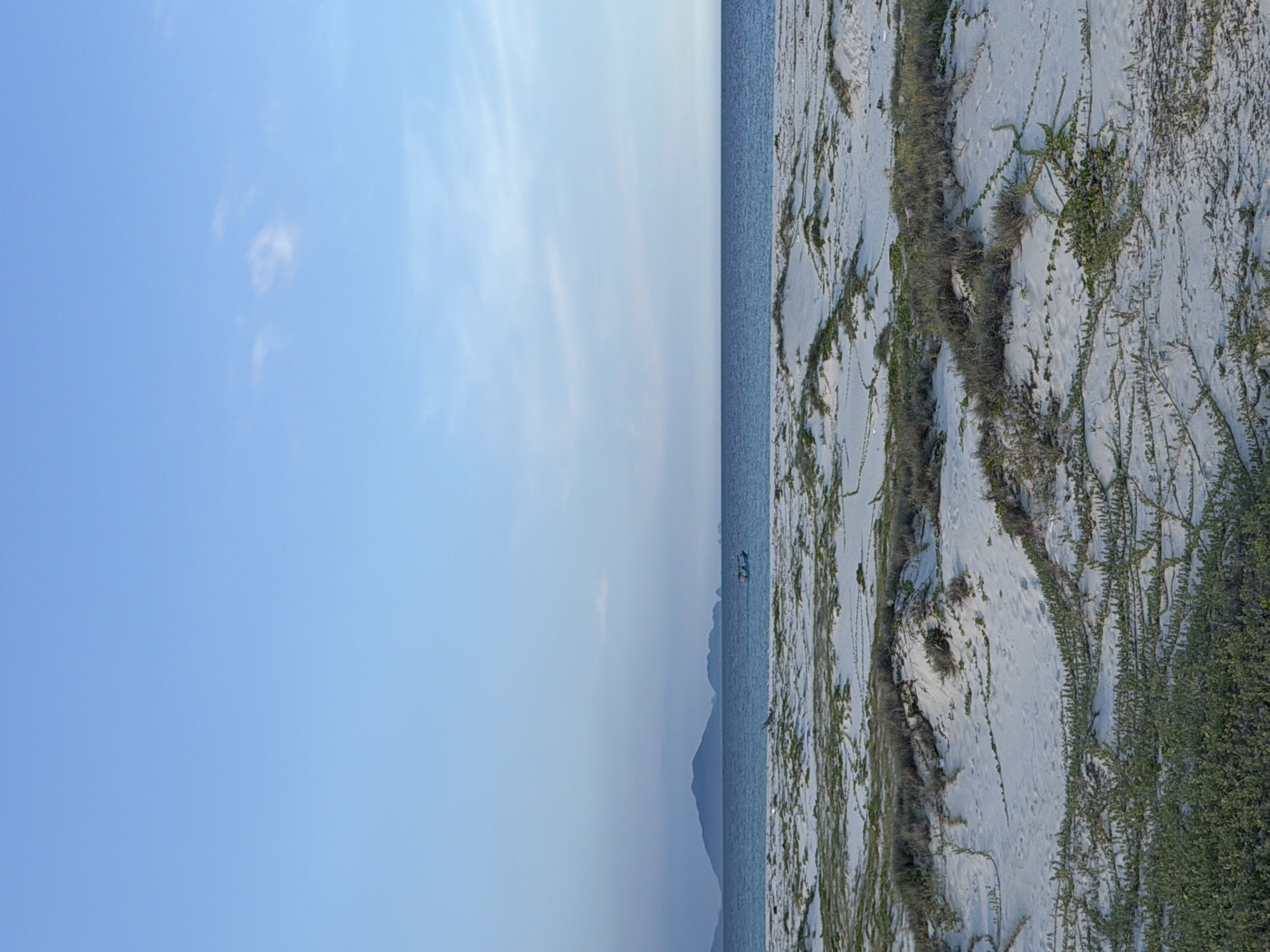 The image depicts a serene coastal landscape in Bac Cam Ranh Ward, Vietnam. In the foreground, a vast expanse of white sand dunes is covered with low-lying green vegetation and creeping vines. The sand is textured and uneven, with subtle impressions of animal tracks. Beyond the dunes, a calm, dark blue ocean stretches to the horizon, meeting a pale blue sky with a few wispy white clouds. In the distance, a small, dark-hulled boat with a light-colored roof is visible on the water. Further in the background, silhouetted against the sky, are the soft outlines of mountains or large islands. The lighting suggests it is either early morning or late afternoon, with a gentle, diffused light illuminating the scene.