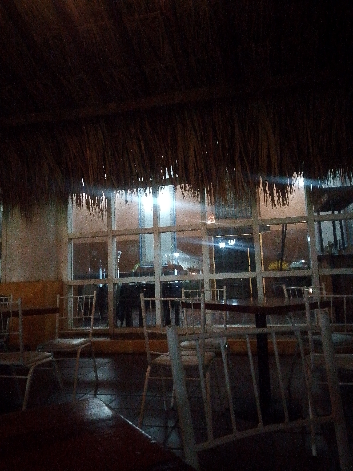 The image shows an outdoor seating area at night, under a thatched roof. The area is furnished with several white metal chairs and dark wooden tables. Large windows reveal the interior of a building, which appears to be a restaurant or bar, with lights on and some bottles visible on shelves. The scene suggests a relaxed, possibly dimly lit, atmosphere. The location is identified as Pedro González, Venezuela.