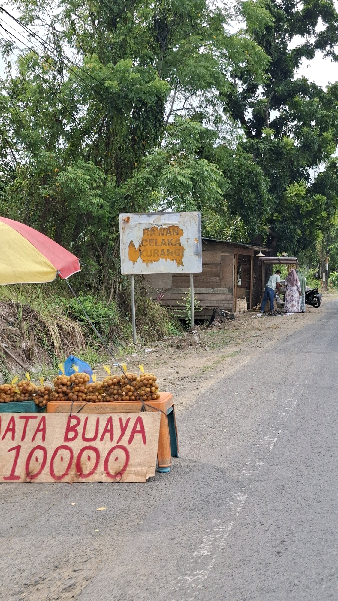 The image depicts a roadside scene in Lhokseumawe, Indonesia, with a focus on a small fruit stand. A yellow and red umbrella provides shade for the stand, which displays bunches of small, brown fruits. A sign in red lettering on a wooden board reads "MATA BUAYA 10000". Behind the fruit stand, a dilapidated wooden shack serves as a small shop or shelter. Two individuals are visible near the shack, one wearing a light green shirt and dark pants, and another in a patterned dress and headscarf. A motorcycle is parked to the right of the shack. In the background, lush green trees and vegetation line the roadside, suggesting a rural or semi-rural setting within the city. A damaged, rusty sign with yellow text is mounted on a pole, partially obscured by foliage. The road itself is paved with a white line marking the center. The overall impression is that of a modest, informal vendor operating along a local road.