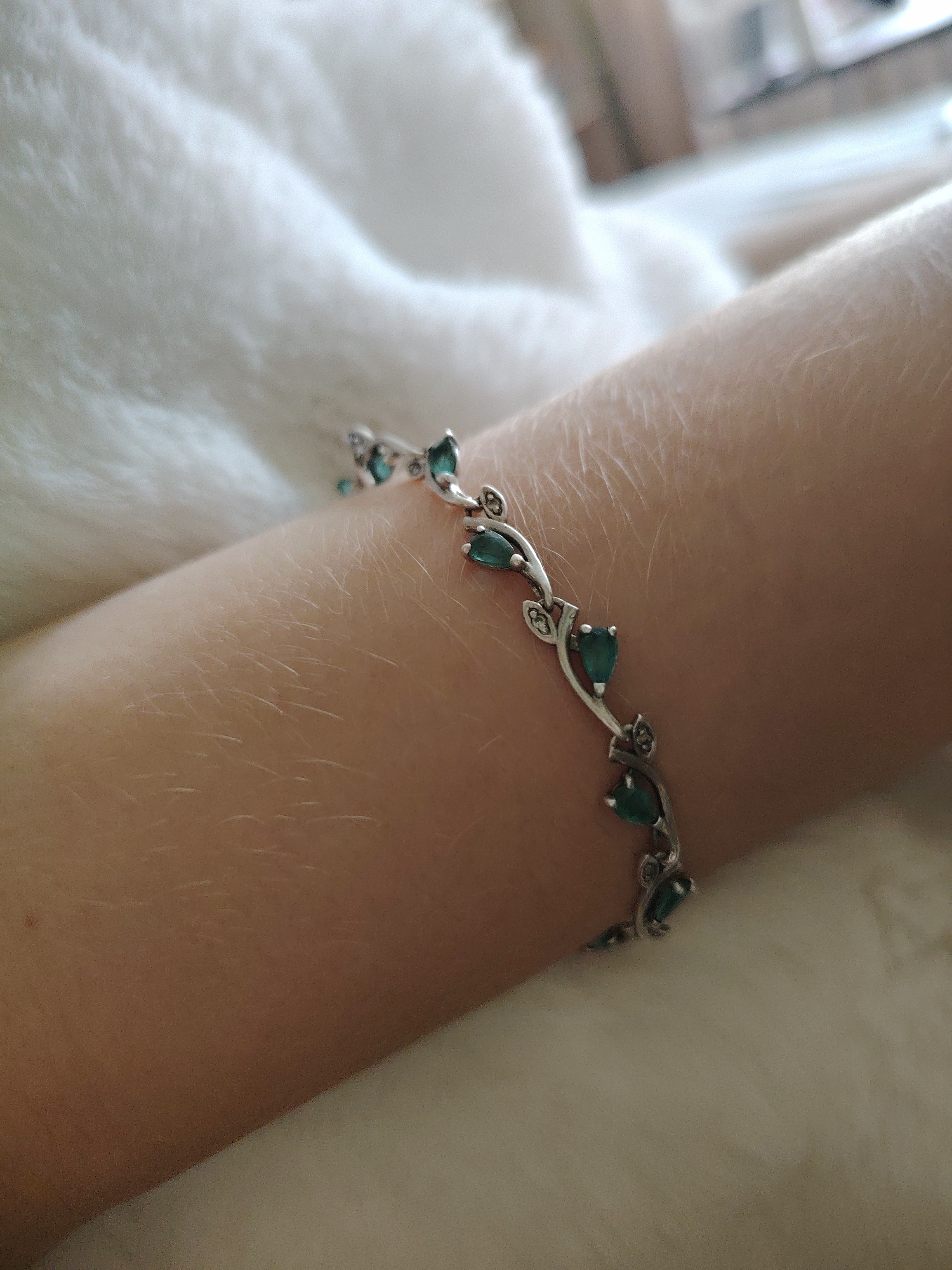 A close-up shot shows a silver bracelet with emerald-colored teardrop-shaped stones and small diamond accents on a person's arm. The bracelet is shaped like a vine with leaves. The arm is covered in fine, light-colored hair and rests on a white, fuzzy surface, possibly a blanket or rug. The background is out of focus and appears to be indoors, with some light-colored furniture visible. The image was taken in an unknown location in Unknown, Unknown.