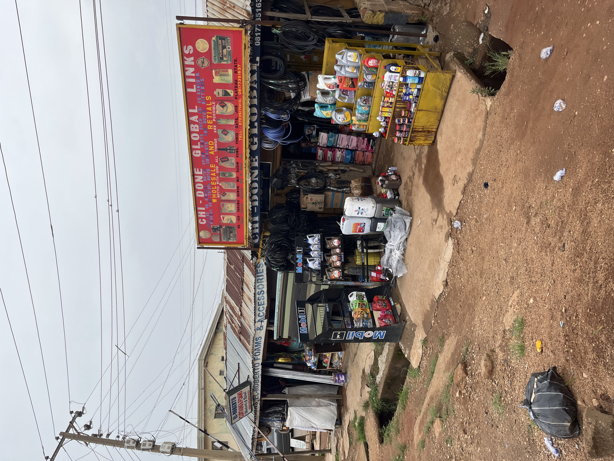 The image presents a street-level view of two commercial establishments in Jos, Nigeria, under an overcast sky. The central focus is "CHI-DONE GLOBAL LINKS," an auto spare parts and accessories shop. A prominent red and white sign for "CHI-DONE GLOBAL LINKS" details its offerings: "MOTOR SPARE PARTS SUCH AS: VOLKSWAGEN, PEUGEOT, TOYOTA, MOTOR LUBRICANTS, AND MOTO BATTERIES." It advertises "WHOLESALE AND RETAILS" with the motto "WITH GOD ALL THINGS ARE POSSIBLE." The sign also provides a physical address: "NO 133 YAKUBU GOWON WAY DADIN KOWA, THRID GATE, JOS PLATEAU STATE" and contact numbers: "07035171010, 08177351637."
Various auto products are displayed both inside the shop and on yellow shelving units outside, including numerous bottles of motor oil (e.g., Mobil 1, POWERLINE Olist), coiled hoses, and what appear to be car batteries and other components. The goods extend onto the unpaved ground in front of the shop.
Adjacent to "CHI-DONE GLOBAL LINKS" on the left is another shop, partially visible, identified by a sign as "EL-ROBERTO CURTAINS & FOAMS & ACCESSORIES," also operating as "WHOLESALE & RETAIL."
The immediate surroundings feature a dirt road with some concrete sections, a visible drainage channel, and scattered litter. Overhead, numerous power lines are strung across the sky, supported by wooden utility poles, indicative of a typical urban or semi-urban street scene. No individuals are visible in the frame. The bright, diffused light suggests it is daytime, likely with heavy cloud cover.