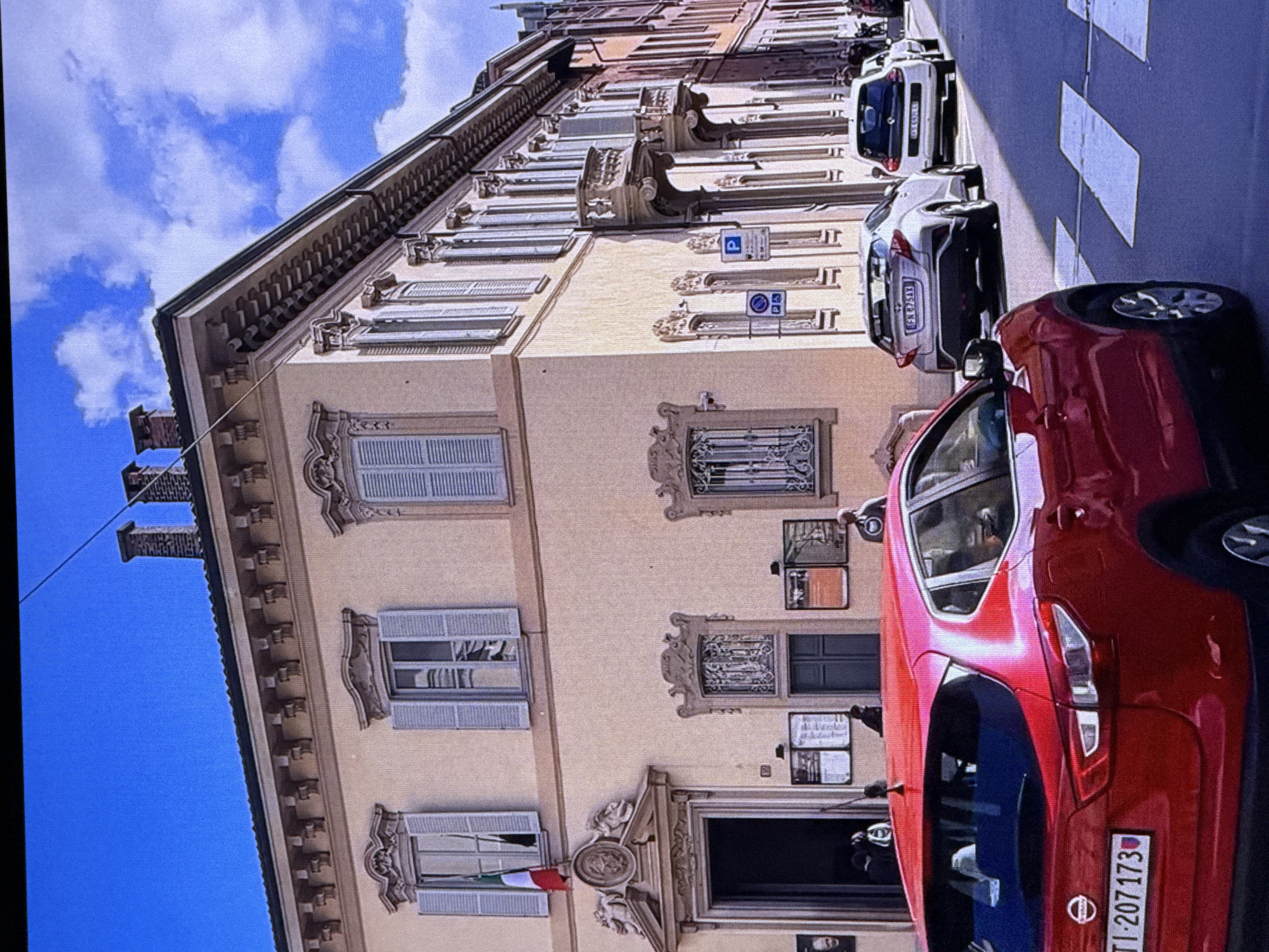 The image shows a street scene in what appears to be an Italian city, not Dubai, United Arab Emirates. A red Nissan Qashqai is parked in the foreground, facing away from the camera. Behind it, a silver Toyota C-HR and a white Renault Twingo are also parked on the street. The buildings lining the street are light-colored and feature ornate architectural details, including balconies and decorative window surrounds. A pedestrian crossing is visible in the lower right of the image. The sky is blue with scattered white clouds. There are a few pedestrians on the sidewalk and in the entrance of one of the buildings. An Italian flag is visible hanging from one of the windows of the building on the left.