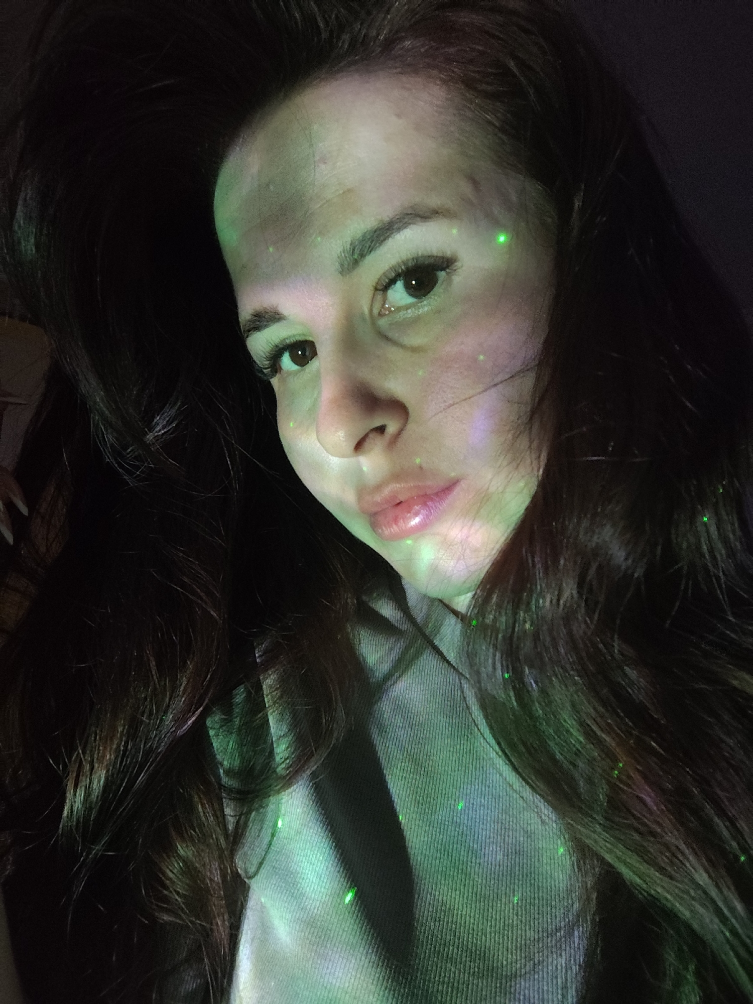 A woman with long, dark hair is photographed in a dimly lit setting, possibly indoors. Her face and upper torso are visible, with green light patterns projected onto her skin and clothing. She is wearing a grey sweatshirt. No specific details indicate a location in Фоки, Russia.