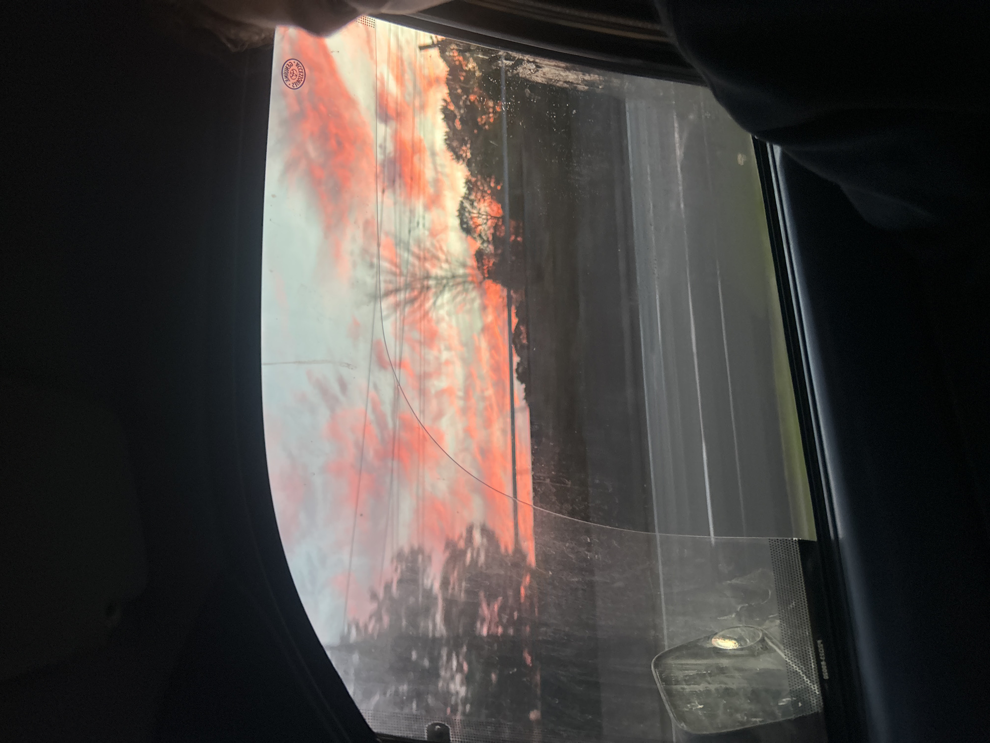 The image captures a sunset viewed from the inside of a vehicle, looking out the passenger window. The sky is dominated by vibrant pink and orange hues, indicative of a sunset. Silhouetted against the colorful sky are trees and a power line. The window itself is slightly dirty and has a curved scratch on its surface. In the foreground, part of the car's interior, including a dark-colored door panel and a person's shoulder and arm, is visible. On the lower left of the window, a part of the side mirror is visible. The scene appears to be on a road, with the suggestion of a highway or similar roadway in the middle ground. Based on the provided location of Hazelbrook, Australia, it is plausible this image was taken in or around that area, given the natural landscape and sunset colors typical of the region.
