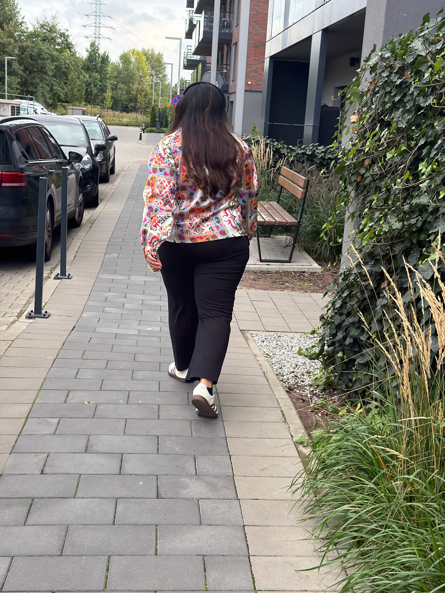 A woman with long, dark hair is walking away from the camera on a paved sidewalk. She is wearing a colorful, patterned button-up shirt, black pants, and white and black sneakers. She is also wearing headphones. The sidewalk is lined with gray pavers and bordered by a small strip of grass and then gravel. To the left of the sidewalk are several parked cars, including a black Audi. In the background, there are trees, a modern apartment building, and a large electrical pylon. A wooden bench is visible on the right side of the image, partially obscured by ivy. The scene appears to be a residential area in the city of Wrocław, Poland.