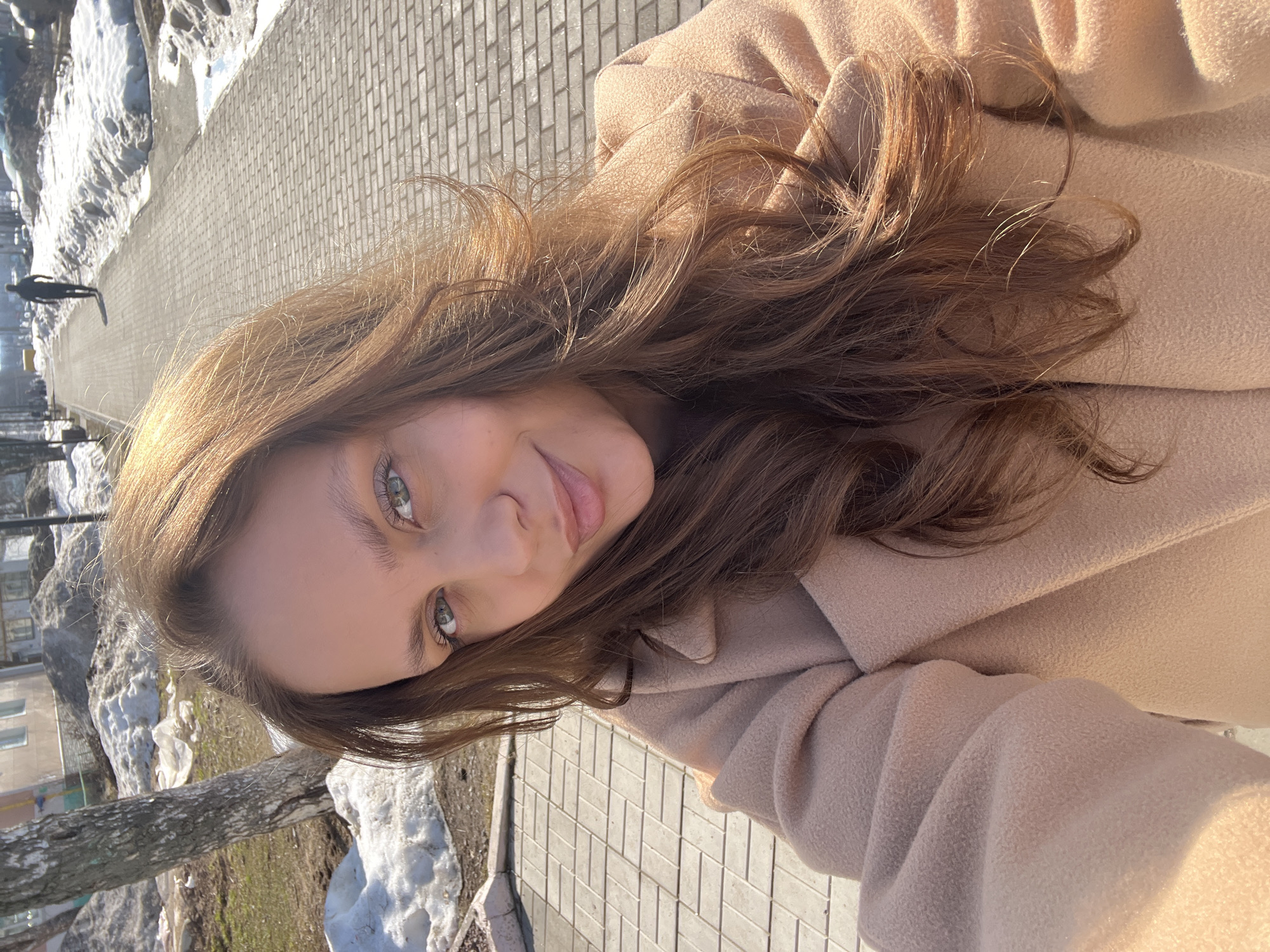 The image depicts an outdoor scene in Chaikovsky, Russia, during clear, sunny weather, indicative of late winter or early spring conditions. A young adult female occupies the foreground, presenting a direct gaze and a subtle, closed-lip smile. She has long, wavy reddish-brown hair and light-colored eyes, and wears a light beige long coat. Strong sunlight illuminates her hair and the right side of her face and coat.
Behind the female, a paved pedestrian path constructed from rectangular grey pavers extends into the distance. Dirty, melting snow is accumulated in large patches on both sides of the pathway, interspersed with areas of bare soil and small puddles of meltwater. A bare tree with light-colored bark is visible on the left side of the path. In the mid-ground, a dark-clad individual walks away from the viewer along the paved path. Further in the background, multi-story residential buildings are discernible on the far left, while a faint yellow object is visible on the right horizon.