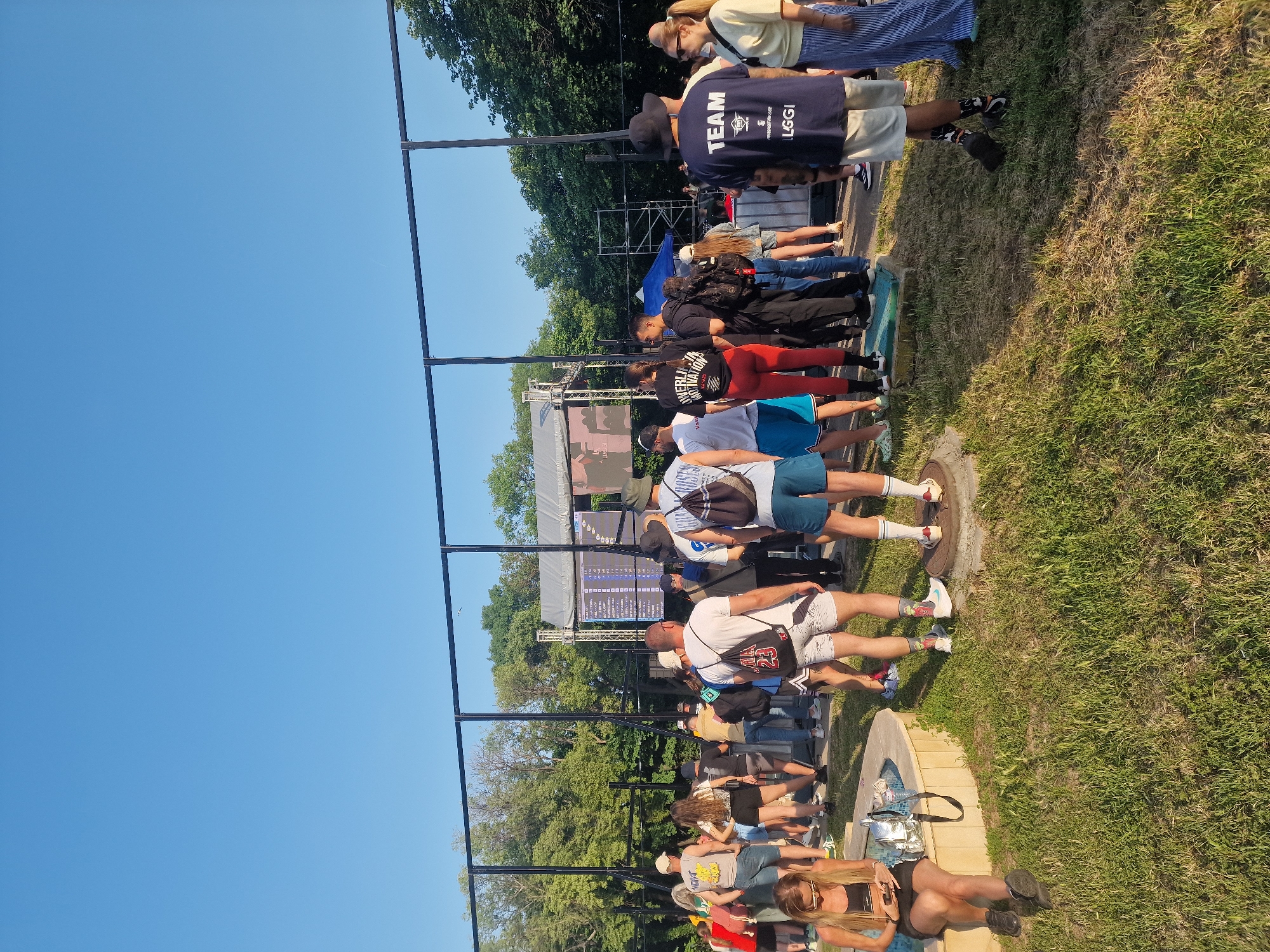 The image captures an outdoor event under a clear blue sky. A crowd of casually dressed individuals, predominantly adults, stands on a grassy area, largely facing two large digital screens positioned behind a tall, temporary black metal fence. The left screen displays white text, possibly scores or data, against a dark background, while the right screen shows a dark image or video. Behind the screens, scaffolding structures and what appears to be a stage are visible, along with a blue tent further back.
In the foreground, on the left, a woman with long light hair and sunglasses sits on a curved, low stone wall or fountain basin, looking at her mobile phone. A silver bag rests beside her. To the right, several individuals stand on the grass. One man in the center wears a white t-shirt with a black drawstring backpack marked "23" and patterned shorts. To his right, another man wears a navy t-shirt with "TEAM" and "LGGI" printed on it, light shorts, and a dark hat. He converses with a blonde woman to his right, who wears a light top and striped blue and white pants. Other attendees are visible throughout the scene, some wearing shorts and t-shirts, including a woman in red leggings and a black t-shirt with "POWERLIFTING MOTIVATION" text on the back. The ground is a mix of green and drier brown grass, with visible metal manhole covers. Lush green trees line the background above the event structures.