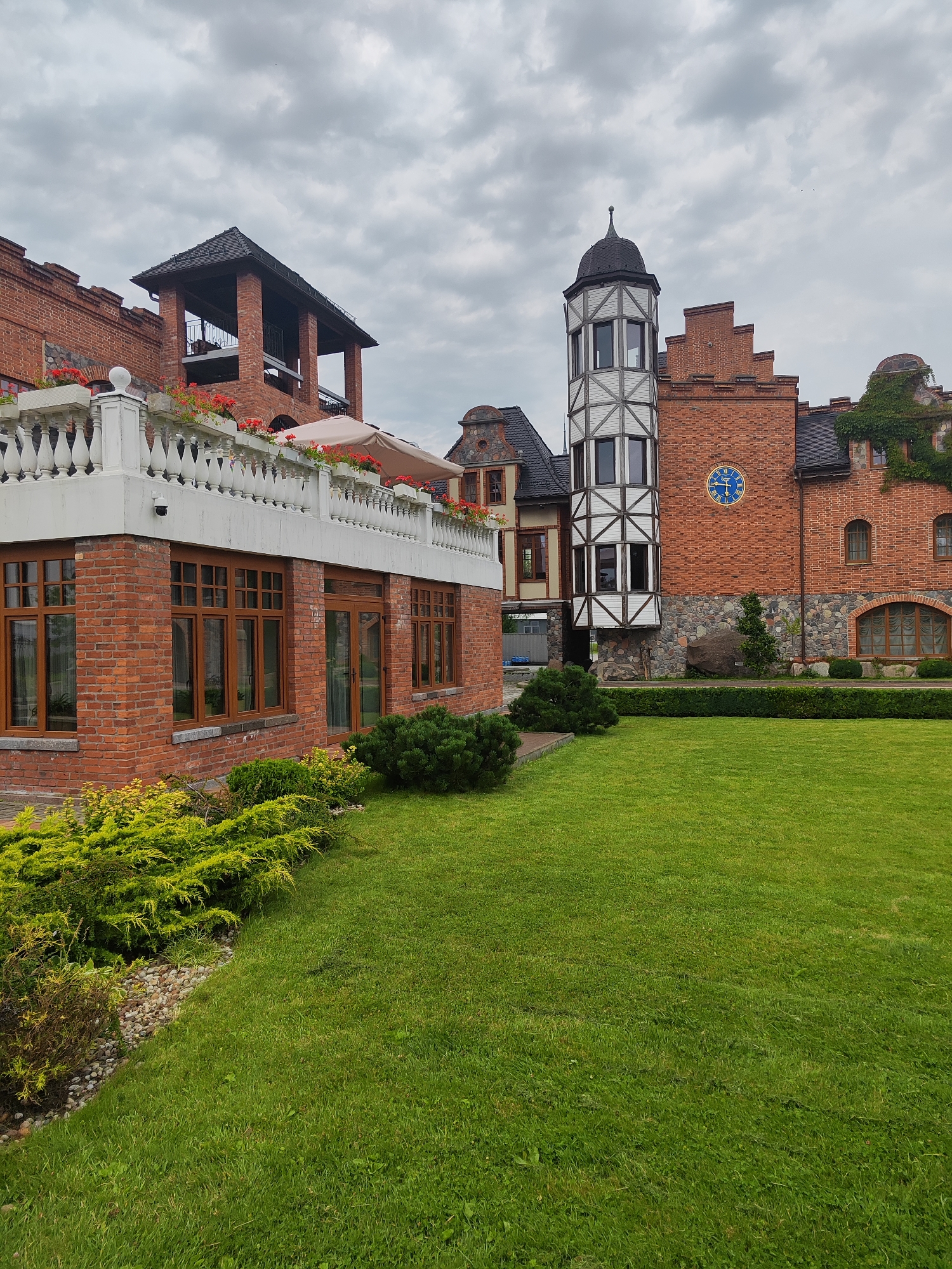 The image shows a multi-story brick building with a European-style architectural design, resembling a castle or manor. The building features a white balustrade on an upper level, a covered balcony, a prominent clock on the facade, and a distinctive turret. The facade is made of red brick with stone accents near the base. The building is surrounded by a well-maintained green lawn and lush vegetation, including hedges and ornamental bushes. The sky is overcast with gray clouds. The image appears to have been taken outdoors during the daytime. The location of this building is in the city of Kaliningrad, Russia, specifically it is the "Ostrov" restaurant complex.