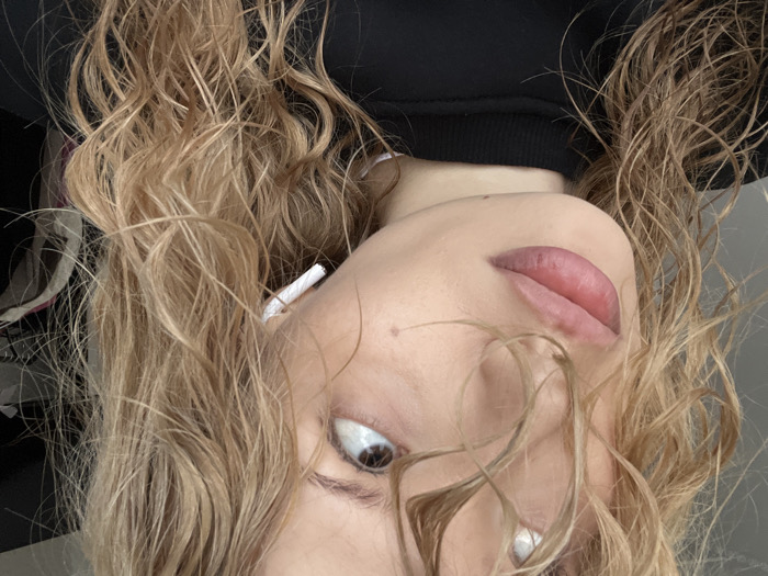 A close-up portrait shot features the left side of a young adult female's face. Her light blonde, wavy hair, appearing wet or styled as such, frames her face, with strands falling across her forehead. Her left eye, featuring a brown iris, is directed upwards and to the left. Her lips are full, slightly parted, and display a pinkish-mauve tint. A small mole is visible on her lower left cheek or chin area. A white wireless earbud is inserted into her left ear. She is attired in a black crew-neck garment. The background is out of focus, consisting of a lighter, indistinct area to the upper left, possibly a wall or window, and a darker, cluttered area to the right, suggesting an indoor environment. The specific location within the city of Chaikovsky, Russia, cannot be determined from the provided image.