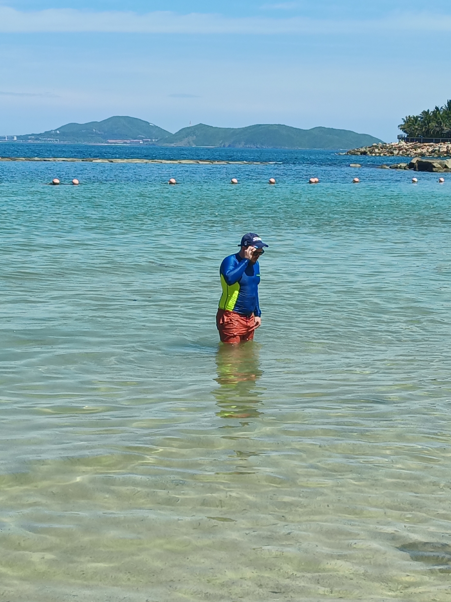 A person stands in clear, shallow water, appearing to adjust a blue baseball cap with sunglasses. They are wearing a blue and yellow long-sleeved rash guard and orange swim trunks. The water is calm with gentle ripples, and the sandy bottom is visible. In the background, a calm blue ocean stretches to a horizon with green, hilly islands under a bright, partly cloudy sky. Orange buoys are visible in a line across the water in the mid-ground.