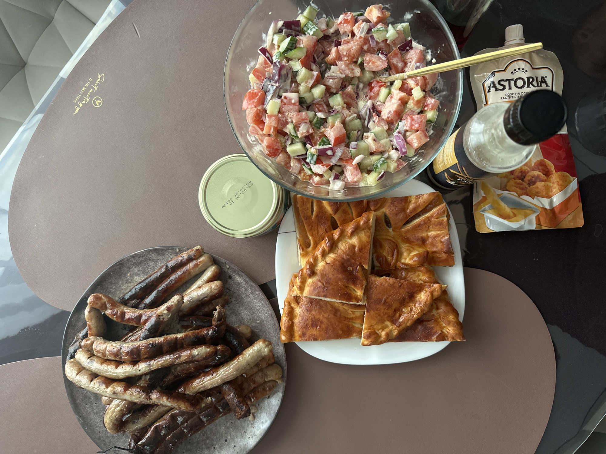 A high-angle shot reveals a table set with a variety of foods, possibly in a home or restaurant in Подшивалово, Russia. A gray plate holds a pile of grilled sausages, some more charred than others. A white plate contains cut pieces of a baked pastry with a golden-brown crust. A clear glass bowl is filled with a salad composed of diced tomatoes, cucumbers, and red onions, dressed with a creamy white sauce, and a gold-colored spoon is placed inside. Nearby is a sealed jar with a green lid, next to bottles and an Astoria brand sauce packet. The tabletop has a brown and black color scheme, with a decorative element displaying the words "Sweet meeting you IS THE BEST THING".