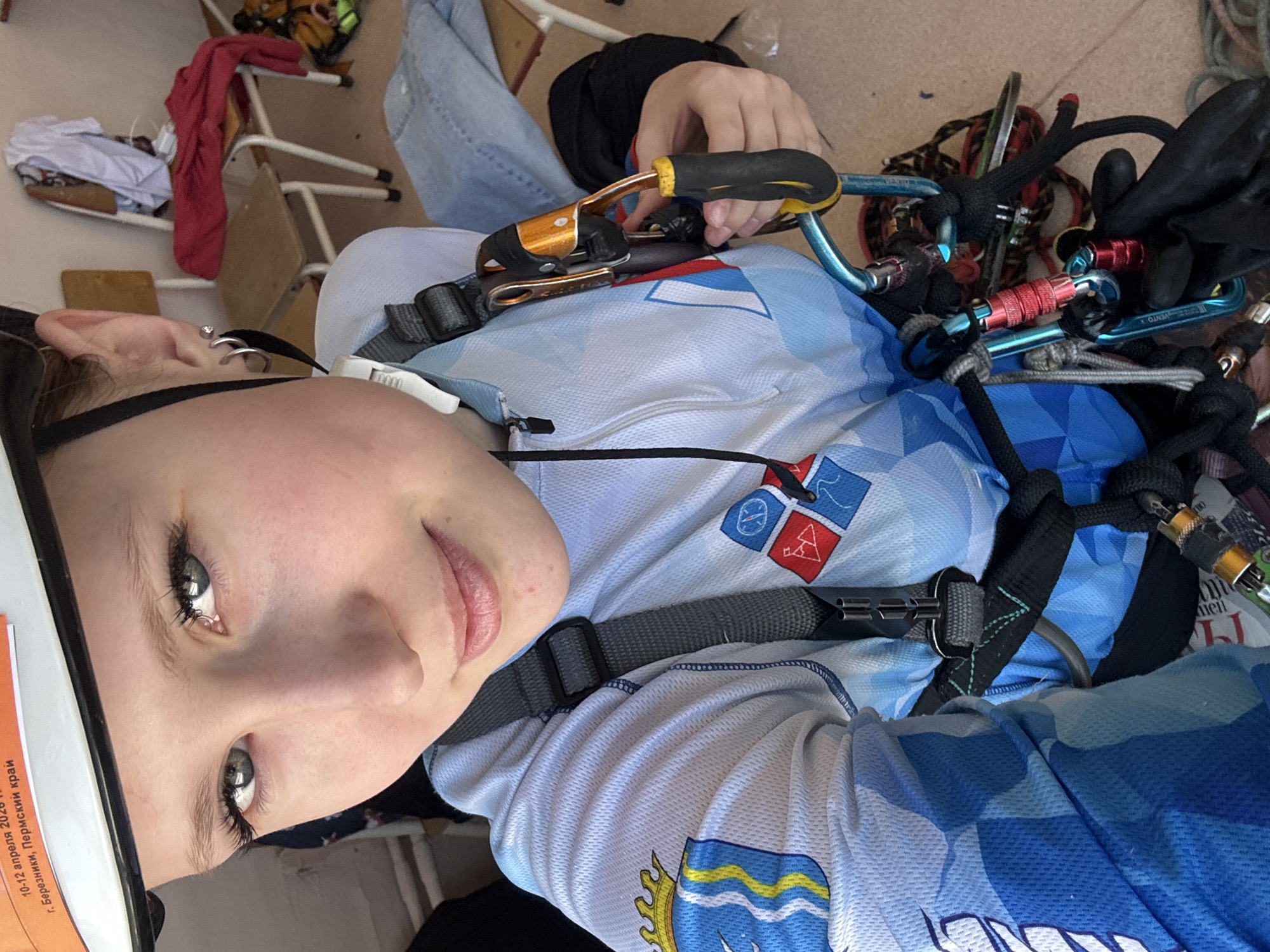 A selfie photograph taken by a Caucasian woman wearing a climbing harness, carabiners, and a helmet. She is wearing a blue and white shirt with logos on the upper left chest and lower abdomen. Text on the helmet indicates the event is in the city of Usolye, Russia. The background is blurred but includes chairs and clothing.