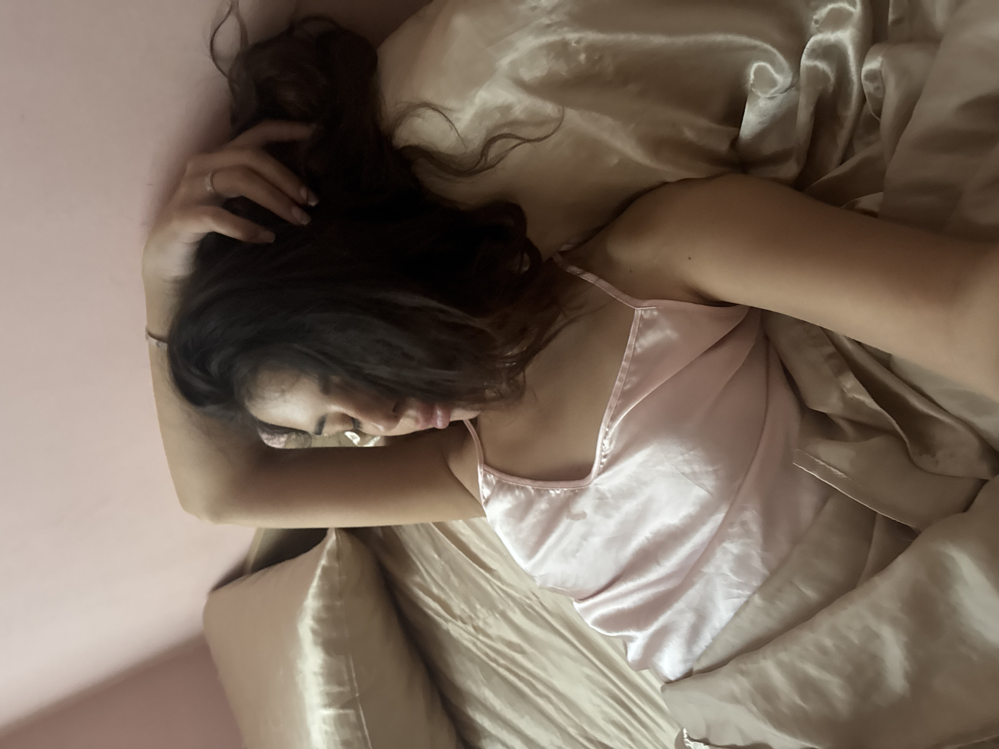 A woman lies in bed, her head resting on a pillow, with her arm bent and her hand near her head. She is wearing a light pink satin slip. The bedding is a neutral satin color. The background wall is painted a pale pink. The scene is indoors, likely a bedroom. There is no discernible information to determine the location in the city of Plovdiv, Bulgaria.