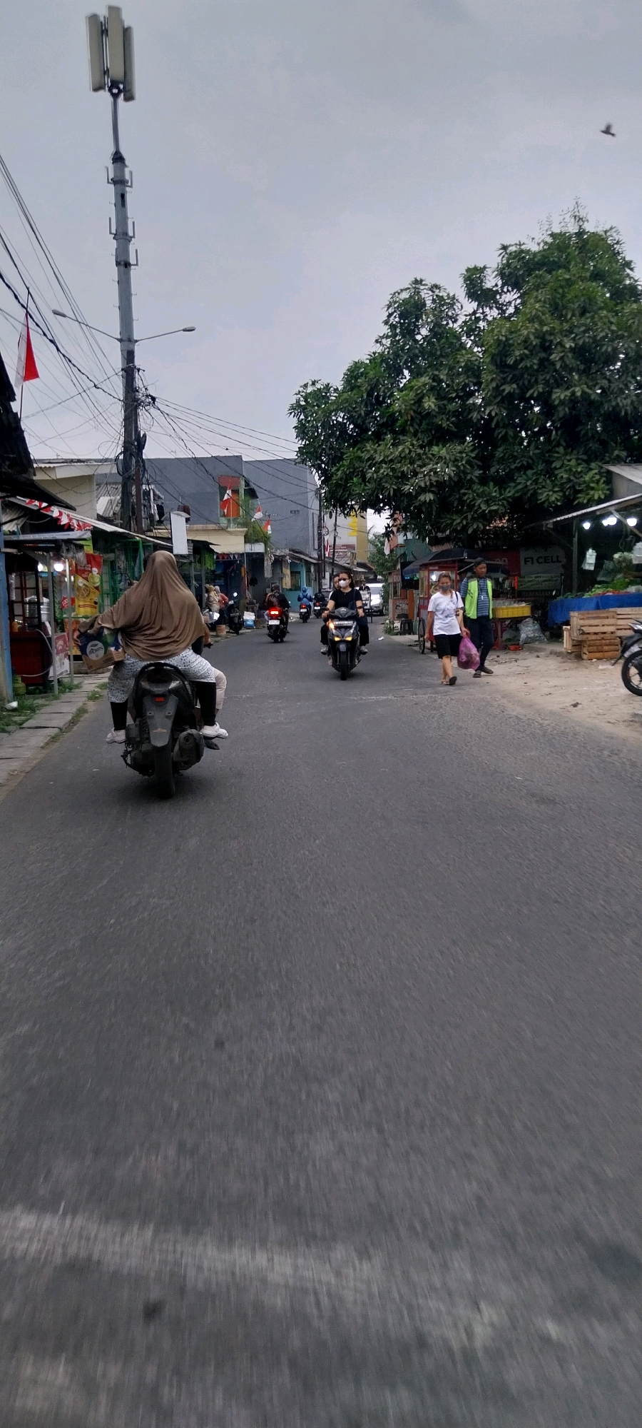 This is a street scene in Kelapa Dua, Indonesia, on a cloudy day. The street is paved and there are buildings on either side. On the left, there are small shops with awnings. On the right, there is a large tree and market stalls selling produce. Several people are present. In the foreground, a person is riding a scooter, wearing a brown hijab and patterned leggings. Further down the street, other individuals are riding motorcycles. Two women are walking on the right side of the street; one is carrying a plastic bag. A cell tower stands on the left side of the road, with wires running from it. Indonesian flags are visible on some of the buildings. The overall impression is of a busy, residential street with commercial activity.