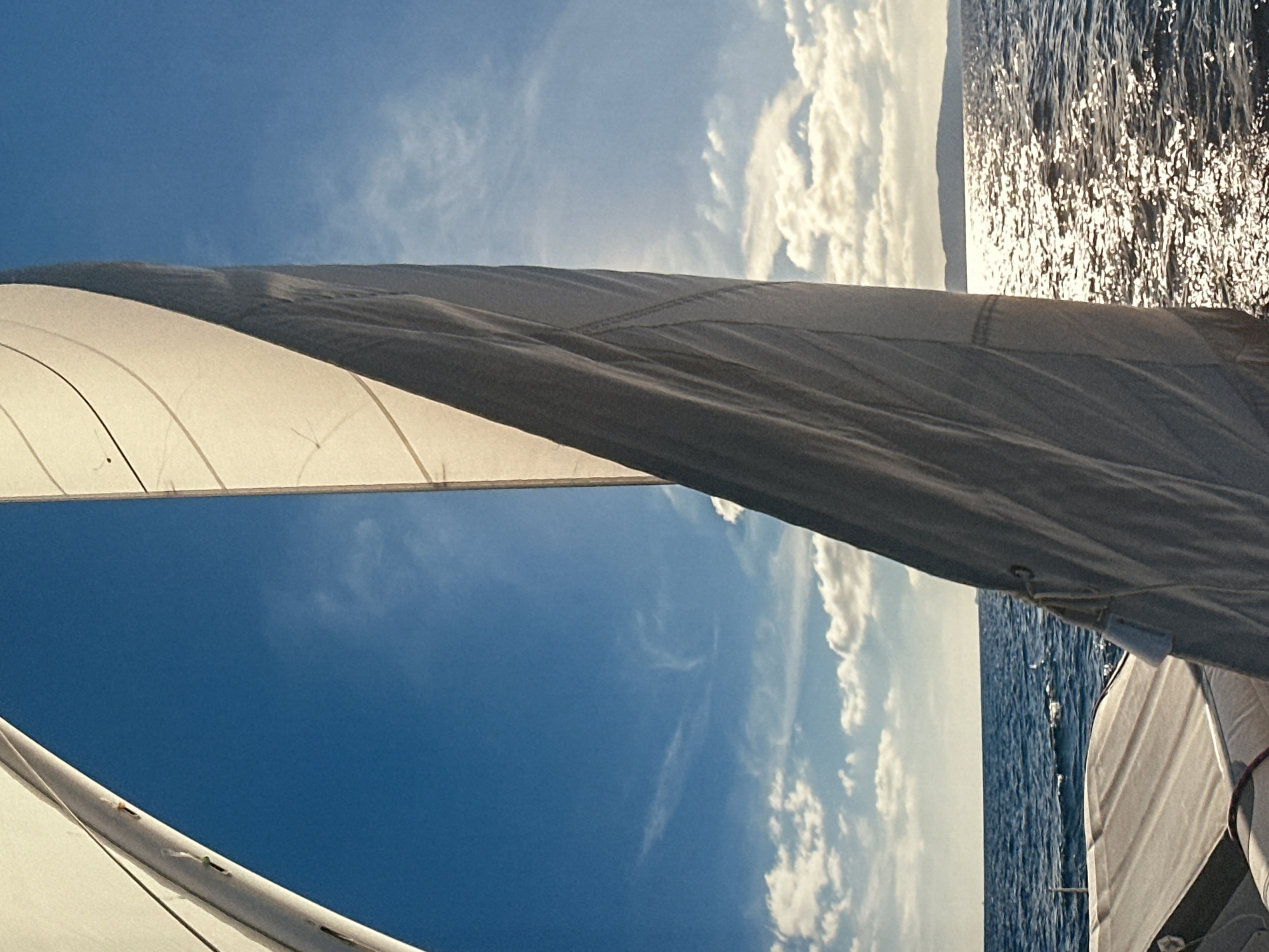 The image is a vertical, low-angle shot from the perspective of someone on a sailboat. Two white sails are prominently featured, one on the left and another larger one dominating the right side of the frame. The sail on the right is catching the sunlight and appears to be billowing. The sky is a vibrant blue with scattered white clouds. The sea is choppy with sunlight glinting off the waves. In the distance, a hazy landmass, possibly an island or coastline, is visible on the horizon. The foreground shows part of the sailboat's deck, including some padded seating. There are no people visible in the image. The scene depicts a bright, sunny day at sea. This image is not of Ossenzijl, Netherlands.