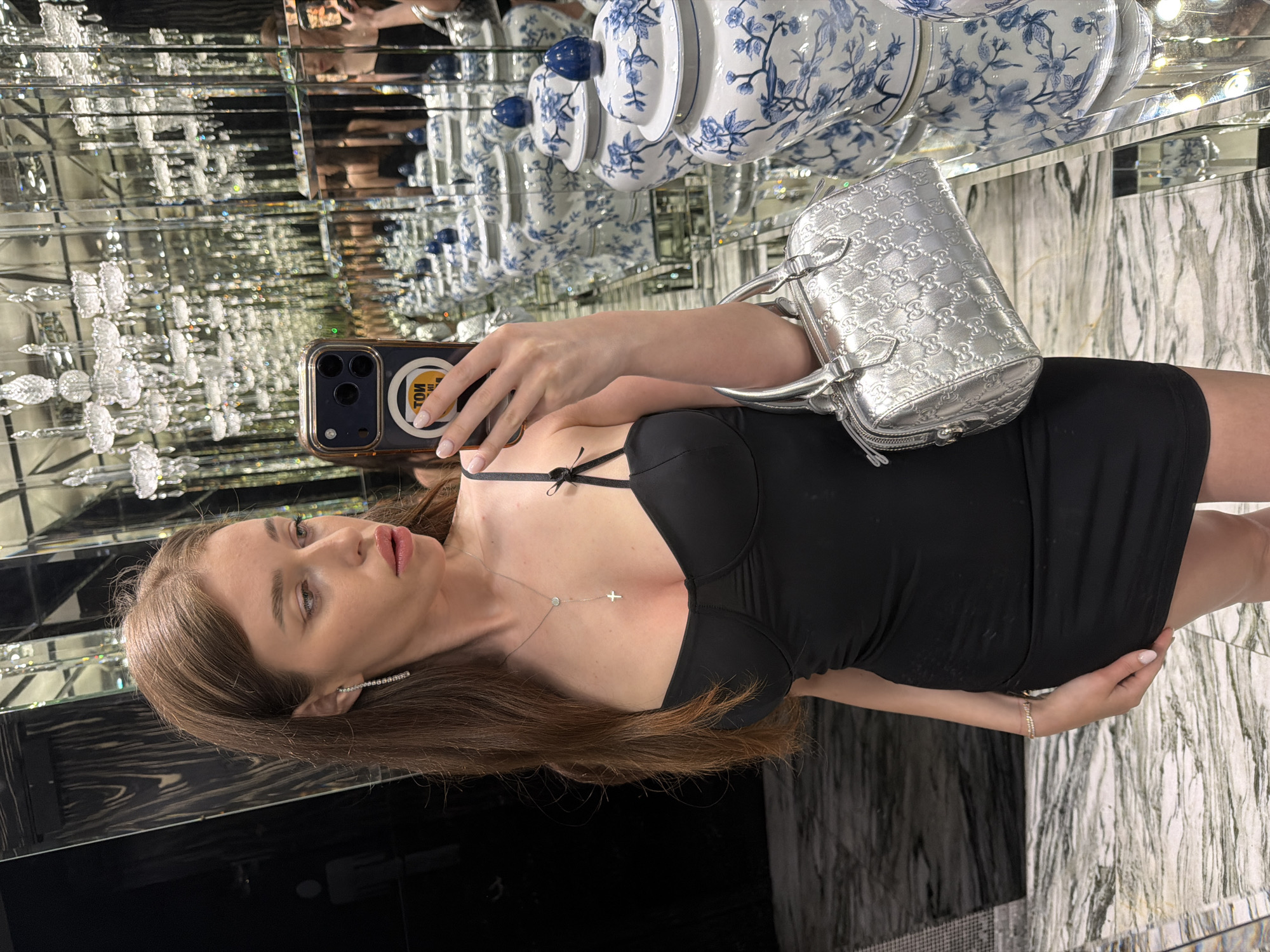 A young adult female is depicted taking a mirror selfie in a highly ornate restroom, identified as being within the Sense Hotel Sofia in Sofia, Bulgaria. The female has long, light brown hair and wears a black, form-fitting mini-dress with thin shoulder straps, one featuring a small bow detail. She is adorned with a long, silver-toned earring on her left ear, a silver-toned necklace with a circular pendant and a cross, and a thin bracelet on her right wrist. Her nails have light-colored polish. In her right hand, she holds a dark-colored smartphone in a clear case, displaying a circular yellow sticker with inverted text, positioned to capture her reflection. Her left hand holds a silver-colored, quilted Gucci handbag with a double-G logo pattern, resting near her hip. The environment features walls entirely covered with rectangular mirror panels, creating an illusion of infinite depth and multiplying reflections of numerous ornate crystal chandeliers that illuminate the space. A prominent display of blue and white Chinese-style porcelain ginger jars with lids, featuring floral motifs, is visible on a mirrored surface that extends throughout the background. The floor is composed of light-colored marble with distinct dark veining. In the far-left background, a dark, reflective panel exhibits a faint, stylized human figure icon, consistent with restroom signage. The subject's gaze is directed slightly downwards and to her right.