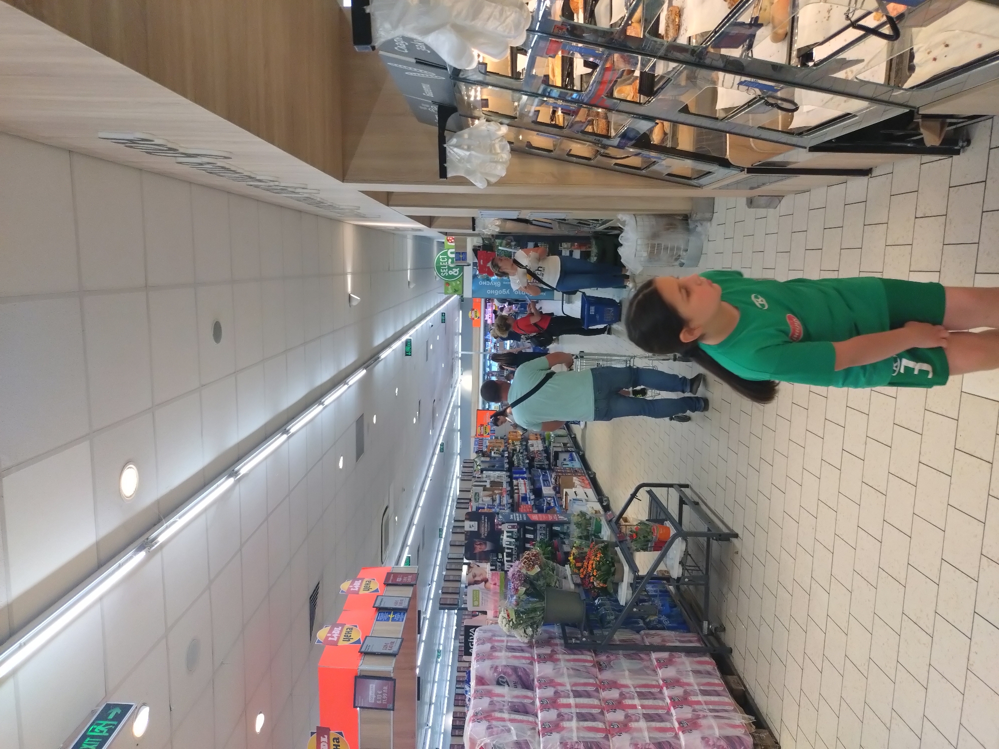 This image, rotated 90 degrees clockwise, captures the interior of a supermarket, likely a Lidl store given the visible branding, in Stara Zagora, Bulgaria. The scene depicts a busy shopping environment with several patrons.
In the immediate foreground, to the right, a young girl with dark hair, wearing a green t-shirt and shorts, stands facing away from the camera, looking towards the left side of the aisle. Her t-shirt has a white logo and the number "3" or "E" in white on the bottom right.
The store features a wide aisle with various product displays. On the left side of the foreground, a large stack of wrapped tissue or toilet paper rolls is visible, next to a display of potted plants and cut flowers. Further down this left aisle, a male shopper in a light blue t-shirt and jeans is seen pushing a shopping cart. The ceiling is white with multiple fluorescent light fixtures running the length of the aisle, along with circular recessed lights. A green "EXIT" sign with an arrow is visible on the ceiling.
To the right, a bakery section with glass display cases showcases various baked goods. White plastic gloves are available for customers, and signs above the display read "Слад" (Sweet), "банички" (banichki, a Bulgarian pastry), and "хляб" (bread).
Further into the background, other shoppers are seen browsing shelves. A green "SELECT" sign is prominent. Red and yellow promotional signs are scattered throughout, typical of a discount supermarket. Visible text on these signs includes "LADL" (Lidl), "цена" (price), and specific prices like "6.13€", "11.99 ЛВ." (Bulgarian Lev), and "129€". Other signs read "Вкусно" (Delicious) and "бързо, удобно" (fast, convenient), "AGIVA", and "SAMA". The floor is laid with light-colored rectangular tiles. The time of day is not discernible, but the bright artificial lighting suggests an indoor daytime or evening setting.