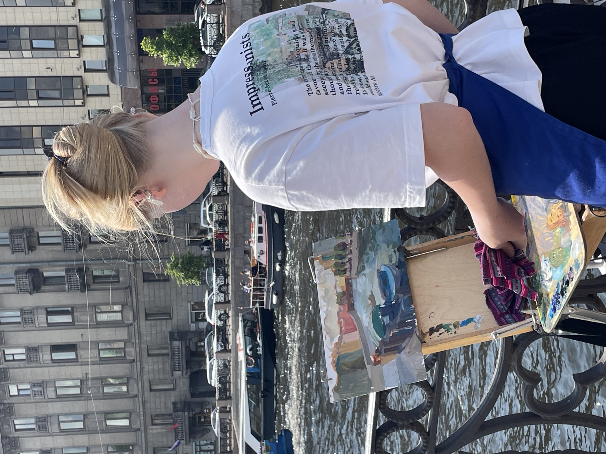 A young person with blonde hair tied in a bun stands on a waterfront in Saint Petersburg, Russia, painting a landscape. The person is wearing a white t-shirt with text and an image on the back, and a blue apron. They are using an easel with a painting in progress, a palette with paints, and a cloth. In the background, there are boats on the river, buildings, and cars. The architecture of the buildings suggests a historical part of the city. The scene is brightly lit, indicating it is daytime.