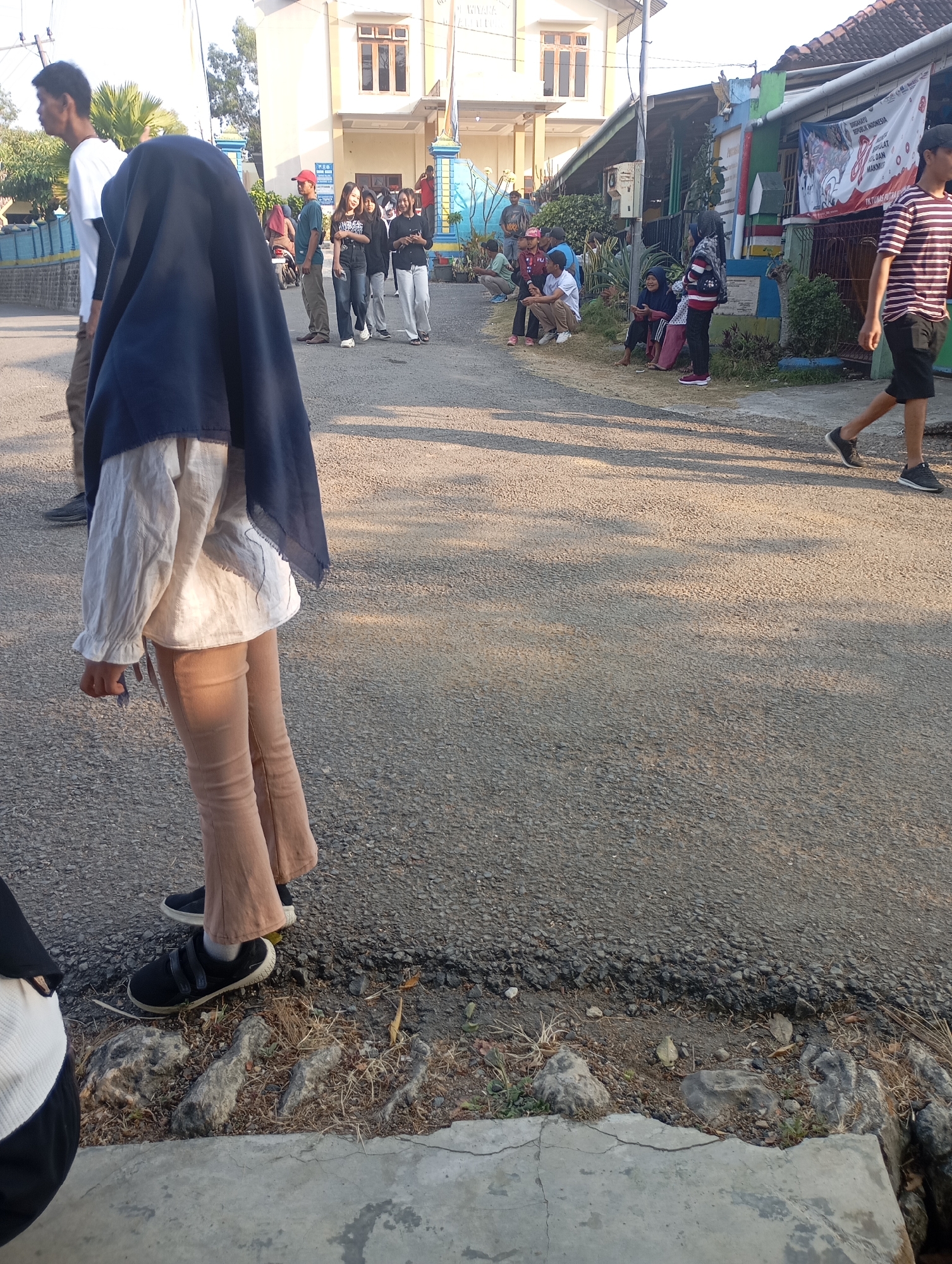In the city of Ketepung, Indonesia, a group of people are gathered on a street in front of a building. The building has a yellow facade with brown trim and multiple windows. To the right of the building is a smaller structure with a tiled roof and a banner displayed.
Several individuals are visible. In the foreground, a person wearing a dark blue hijab, a white long-sleeved shirt, and tan pants is standing and facing away from the camera, with their legs and feet visible. They are wearing black sneakers with velcro straps. To the left of this person, another individual in a white t-shirt is partially visible.
Further down the street, a man in a blue shirt and tan pants is walking. To his right, a group of people are seated or standing near the smaller building. Among them are individuals wearing t-shirts and jeans, some are looking at a banner.
On the right side of the image, a person in a striped t-shirt and dark shorts is walking away from the camera. Shadows of people are cast on the asphalt road. The road is paved with asphalt and has a rough texture. The environment appears to be a sunny day, with the sun casting long shadows. The ground along the side of the road is covered with gravel, dry grass, and rocks.