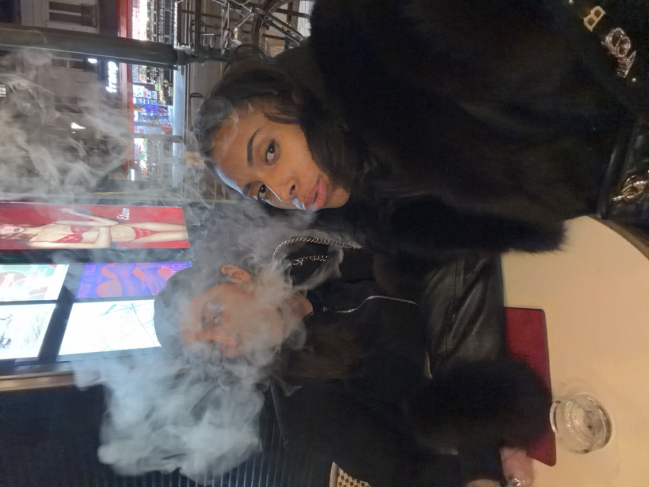 Two women are captured in a selfie at an outdoor location. One woman is blowing out smoke while the other is in the background. Both have dark hair, and are dressed in black. There's an Elam lingerie advertisement displayed in the background. The scene suggests a nightlife setting with bright store signs and urban elements visible.