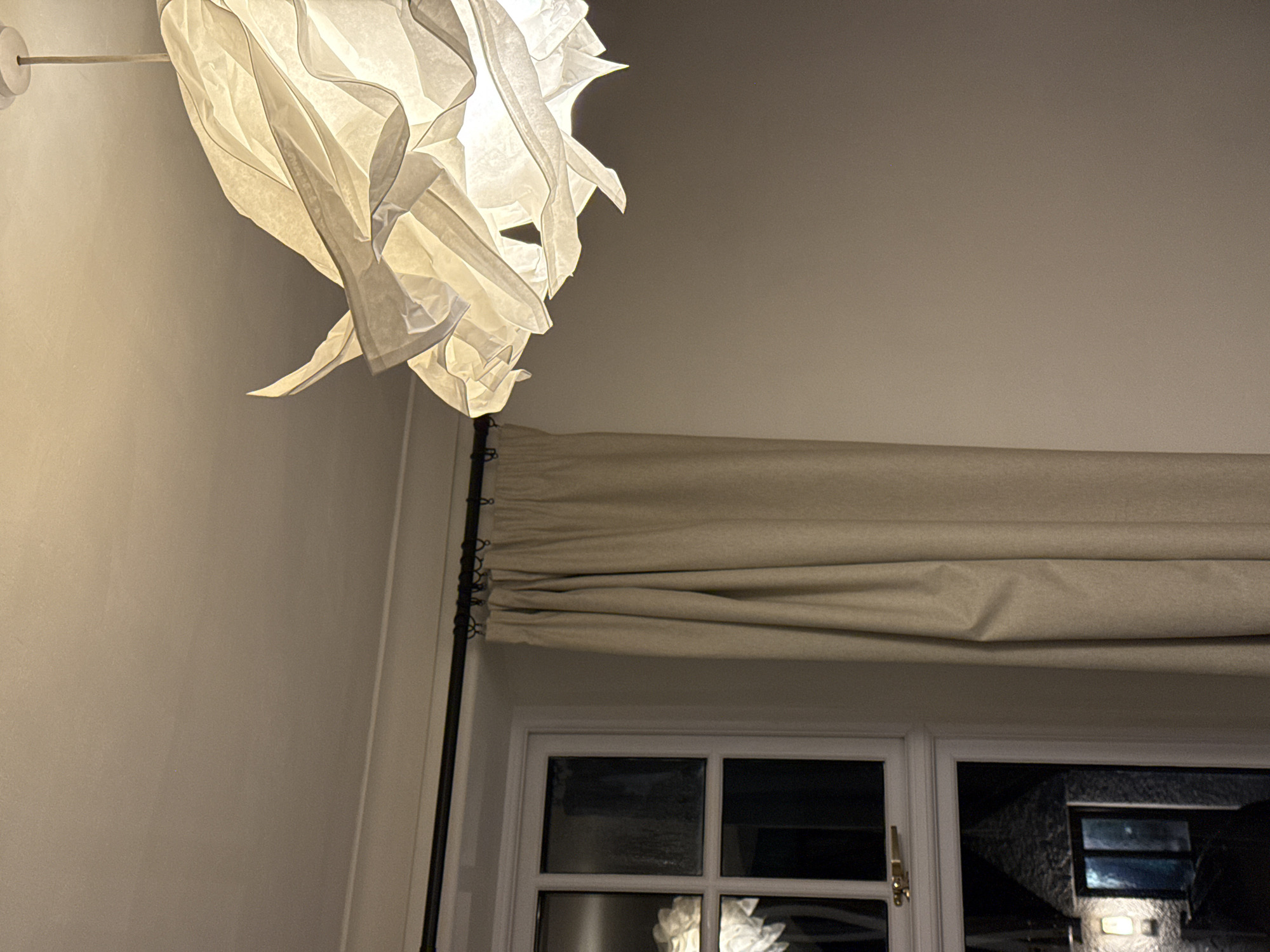 The image presents an interior view under low lighting conditions. Dominating the upper left is an illuminated, stylized paper lantern hanging from the wall. The walls appear to be off-white or light beige. Below the lantern, a dark metal rod extends downwards. To the right, a beige roman blind is positioned above a white-framed window. The window has a multi-pane design, and through it, a dark, indistinct background is visible, possibly an outdoor scene at night. No people are visible in the shot. The location is impossible to determine based on the image.