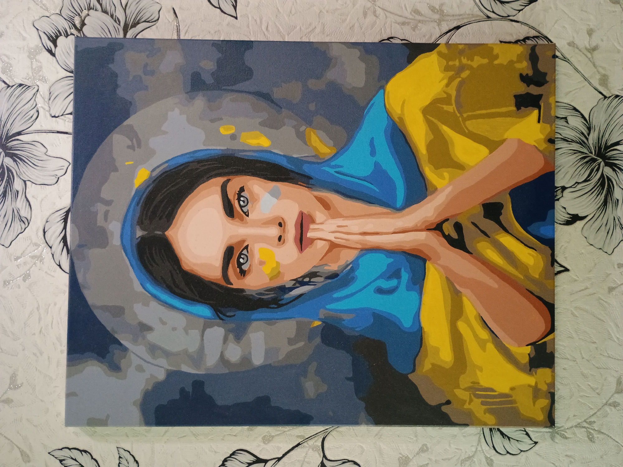 The image shows a painting of a woman with her hands clasped in prayer. She is depicted with a blue and yellow headscarf, reminiscent of the Ukrainian flag. Her eyes are looking directly at the viewer, and there are yellow and white markings on her cheeks. The background is a blend of dark blue, grey, and yellow, with some abstract shapes. The painting appears to be a "paint by numbers" style artwork, with visible outlines and distinct color blocks. The painting is displayed on a wall covered with floral wallpaper. There is no indication of a specific location in the city of Zaporizhzhia, Ukraine.