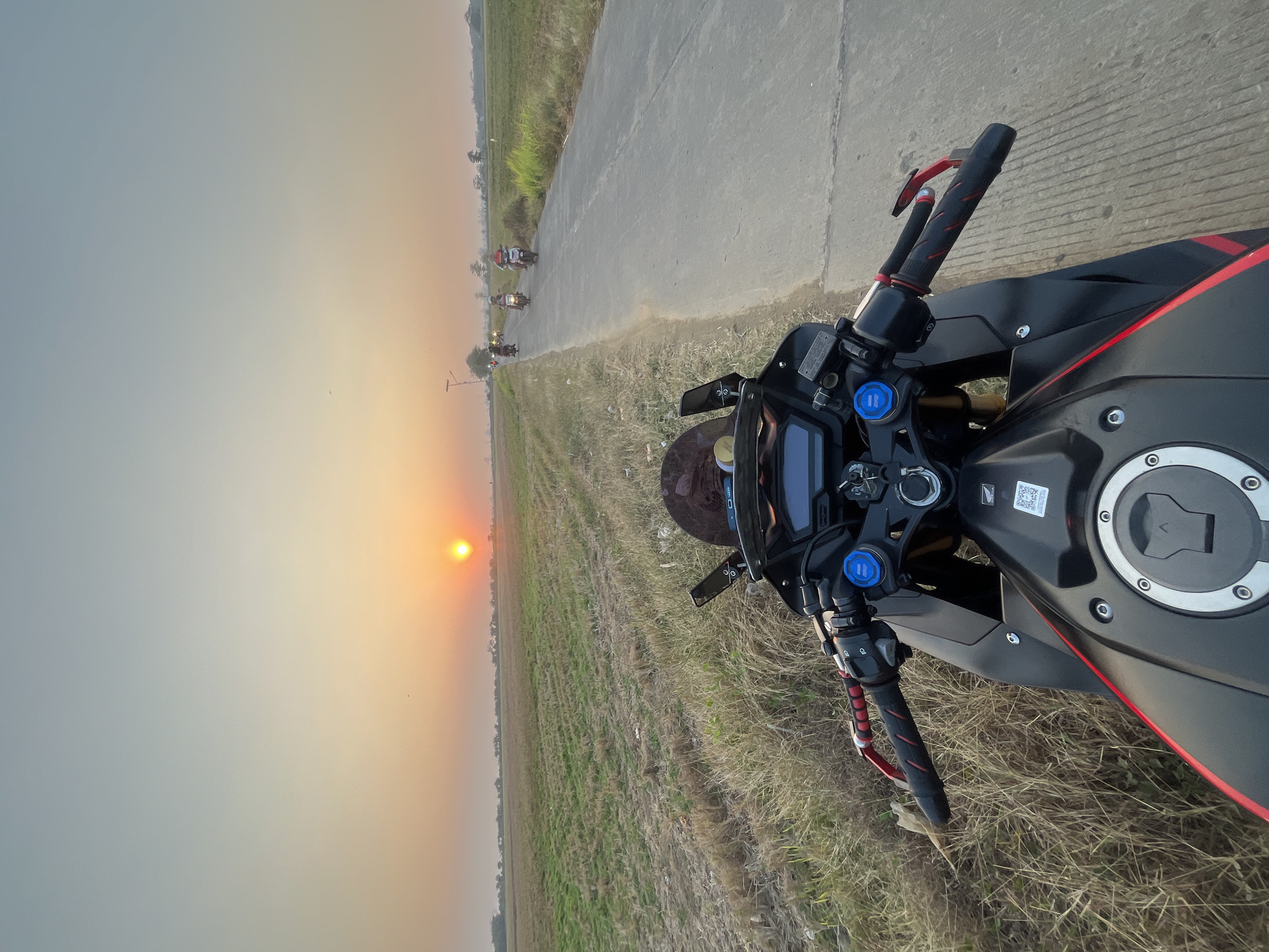 A first-person perspective shot of a black and red motorcycle parked on the side of a rural road in Troso, Indonesia, during sunset. The motorcycle is facing forward, with its handlebars, dashboard, and fuel tank visible in the foreground. The road stretches into the distance, lined with dry grass on the left and a field of green crops on the right. Several other motorcycles are visible further down the road, moving away from the camera. The sun is setting on the horizon, casting a warm orange glow across the sky and illuminating the landscape. The overall scene evokes a sense of tranquility and adventure.